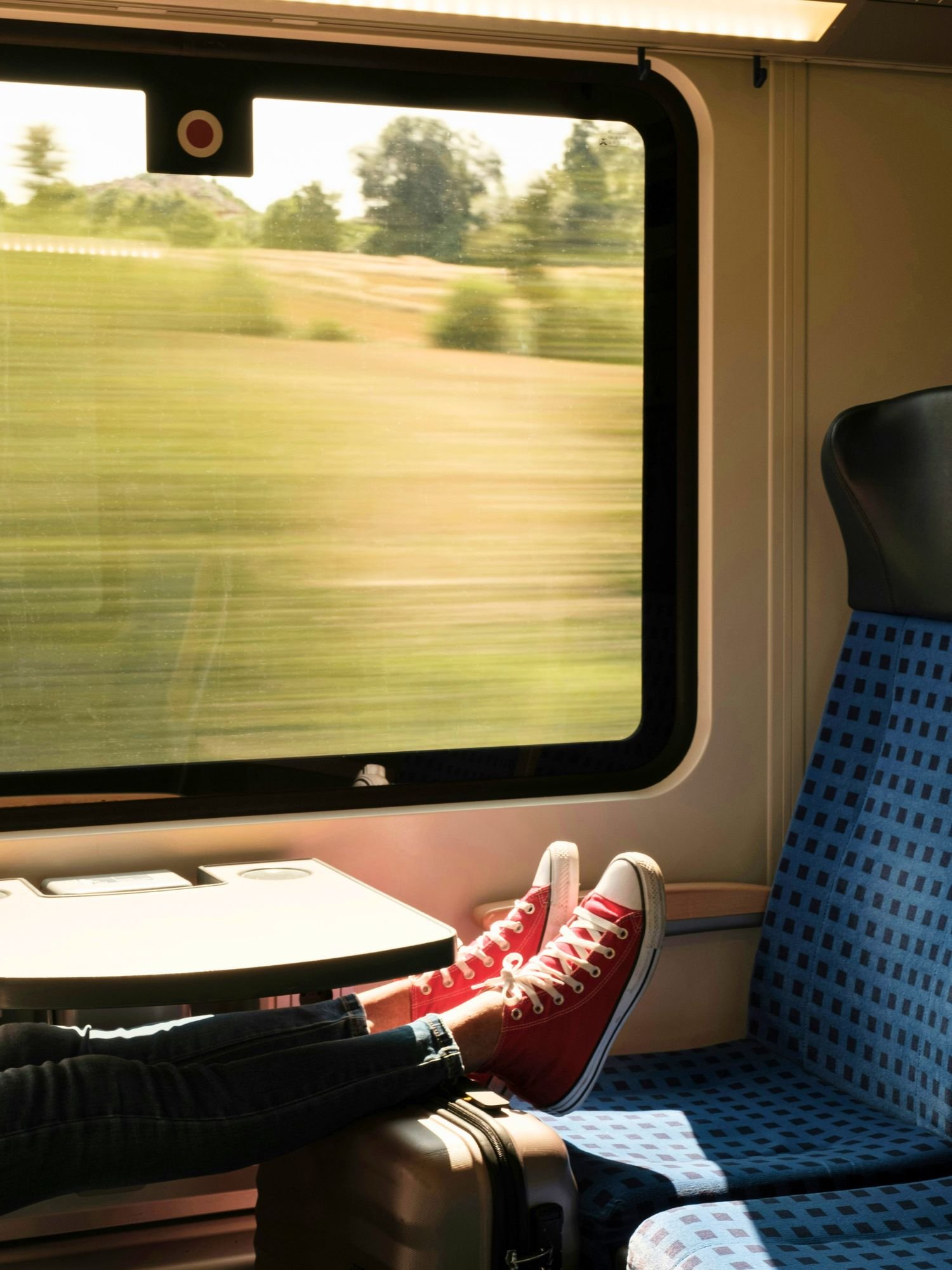 A person’s legs, wearing dark denim jeans and bright red high-top sneakers with white laces, are stretched out inside a moving train, resting their feet on a silver suitcase.