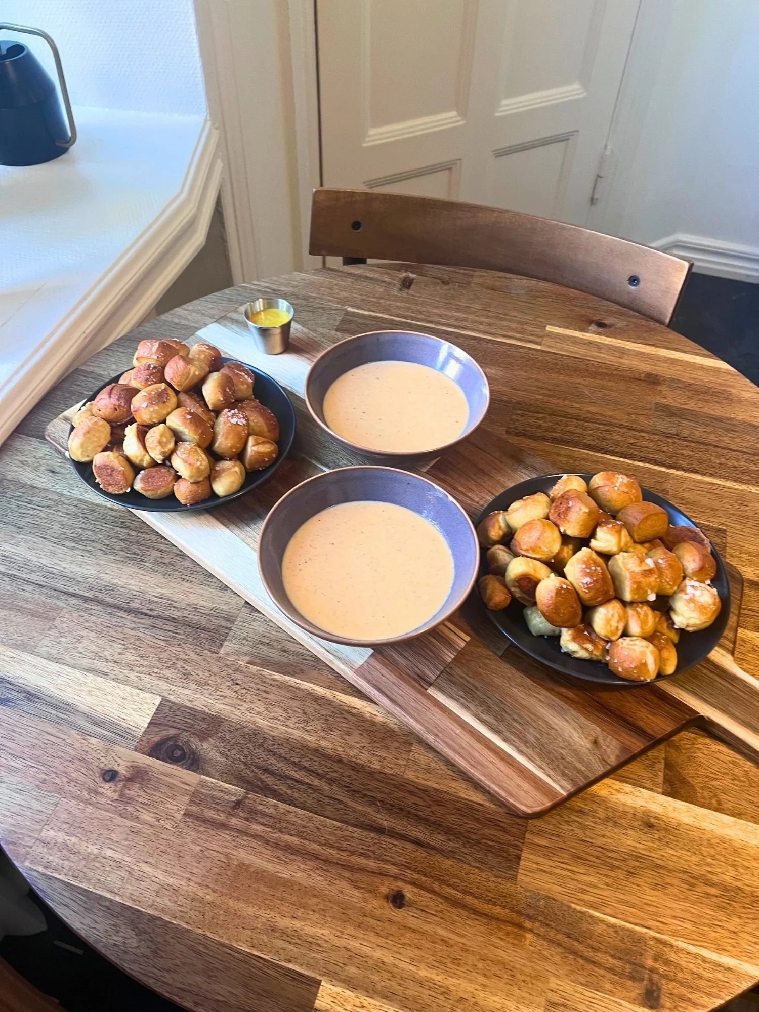 High-angle shot of a wooden serving board set across a round wooden table, holding two plates of pretzel bites, two bowls of cheese dip, and a small metal ramekin of mustard sauce.