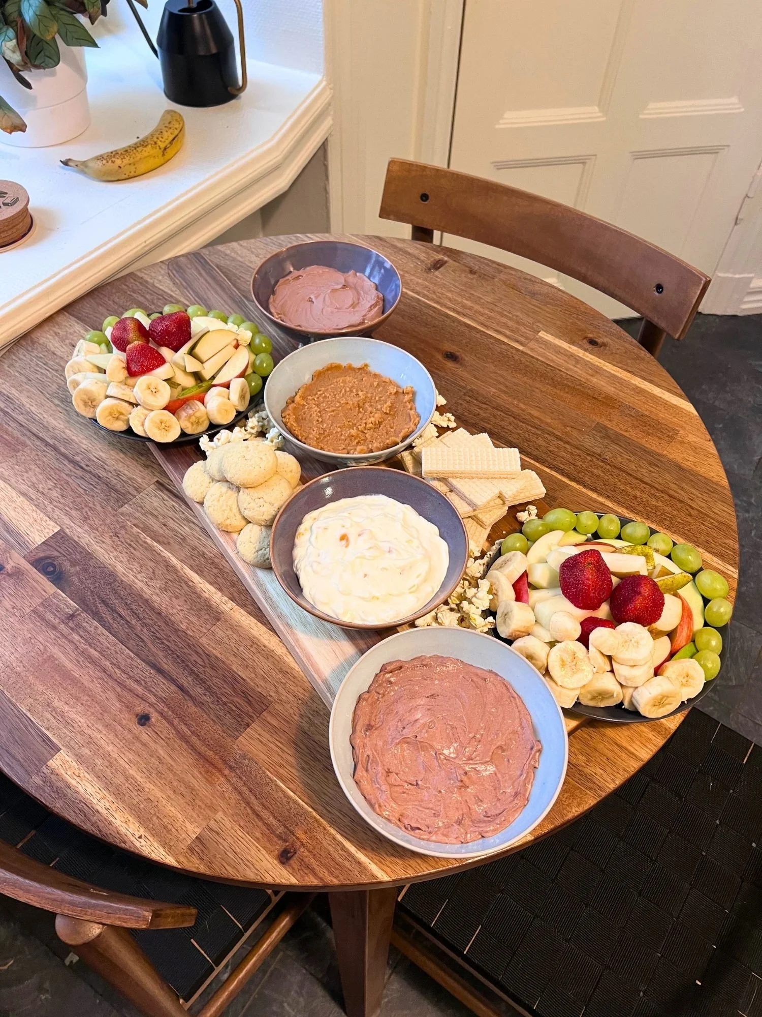 A wooden table set with a serving board filled with two fresh fruit platters, four ceramic bowls of dessert dips, light cookies, stacked wafer biscuits, and popcorn.
