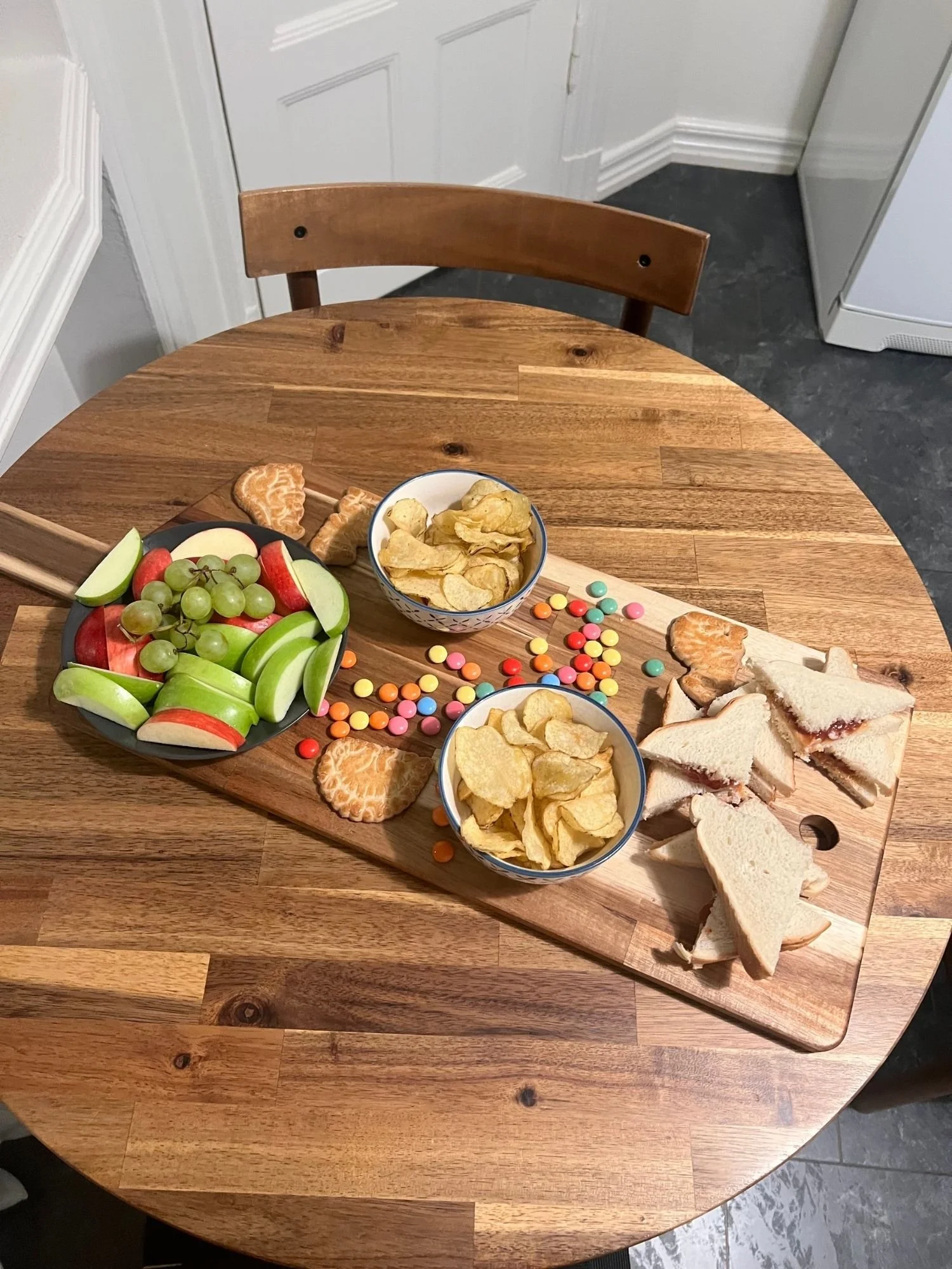 High-angle shot of a rectangular wooden serving board on a round wooden table, set with a child-friendly spread featuring sandwich triangles.