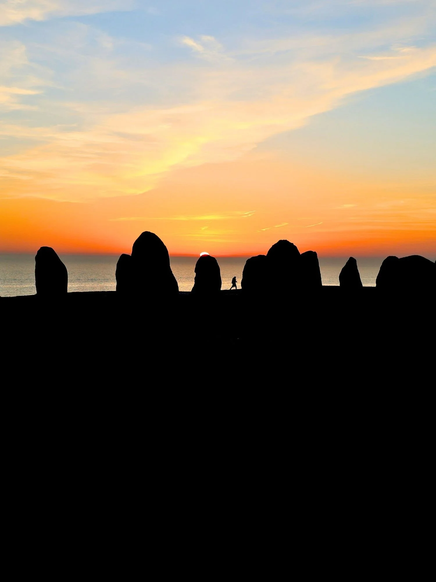 The Ales Stenar megalithic monument at sunset. The ancient standing stones are captured in sharp black silhouette against a vibrant horizon where the sun is just beginning to dip below the sea.