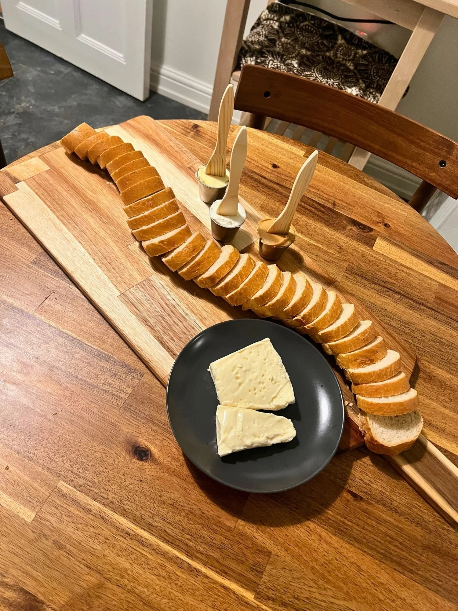 A long wooden serving board on a round wooden table, featuring an S-curved line of sliced baguette, a dark plate with soft cheese, and small ramekins with spreads and wooden knives.