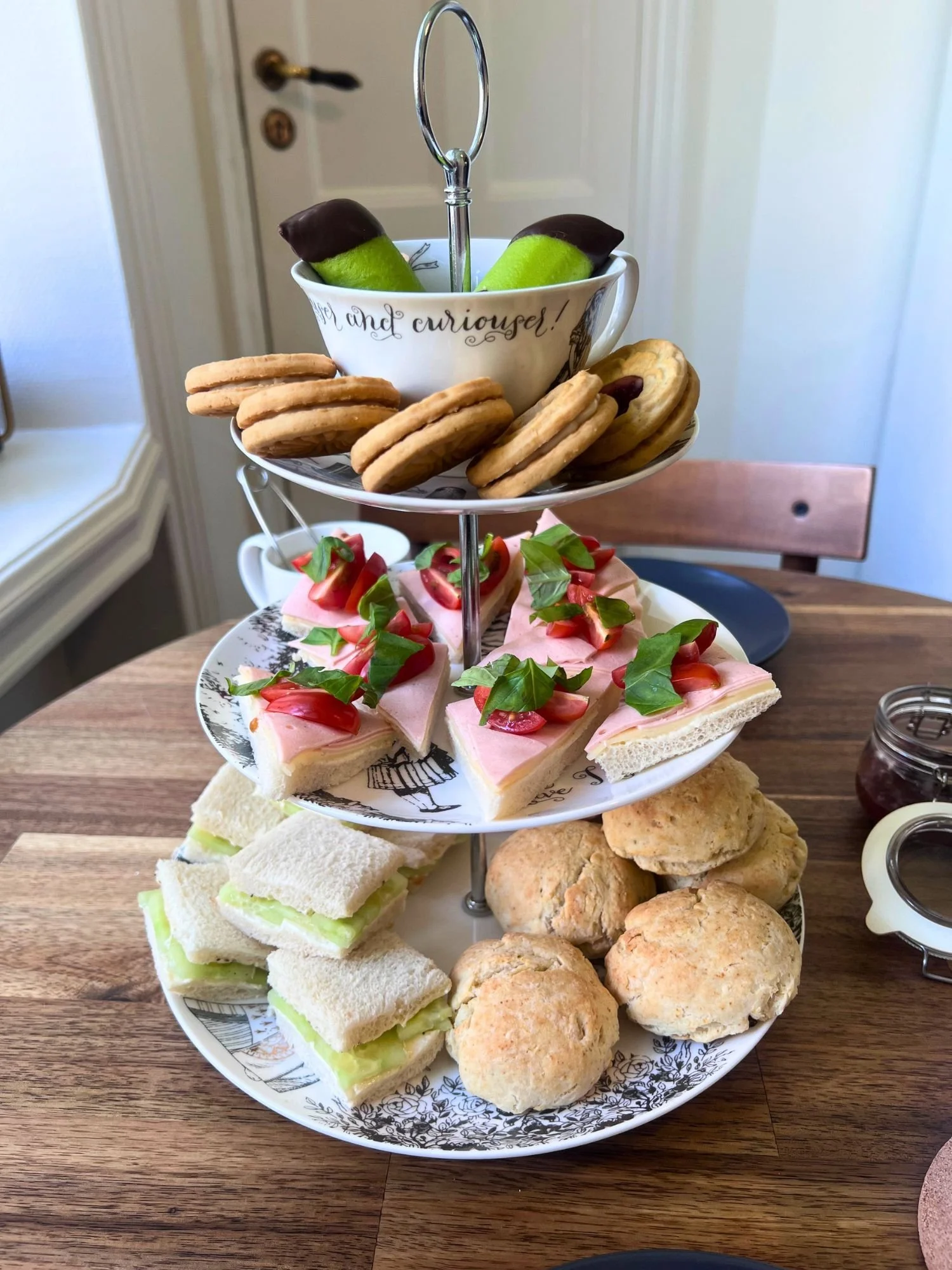 A three-tiered afternoon tea stand on a wooden table. The top tier has a white teacup with black cursive text holding two green marzipan dammsugare pastries with chocolate ends, surrounded by Ballerina sandwich cookies on the saucer.