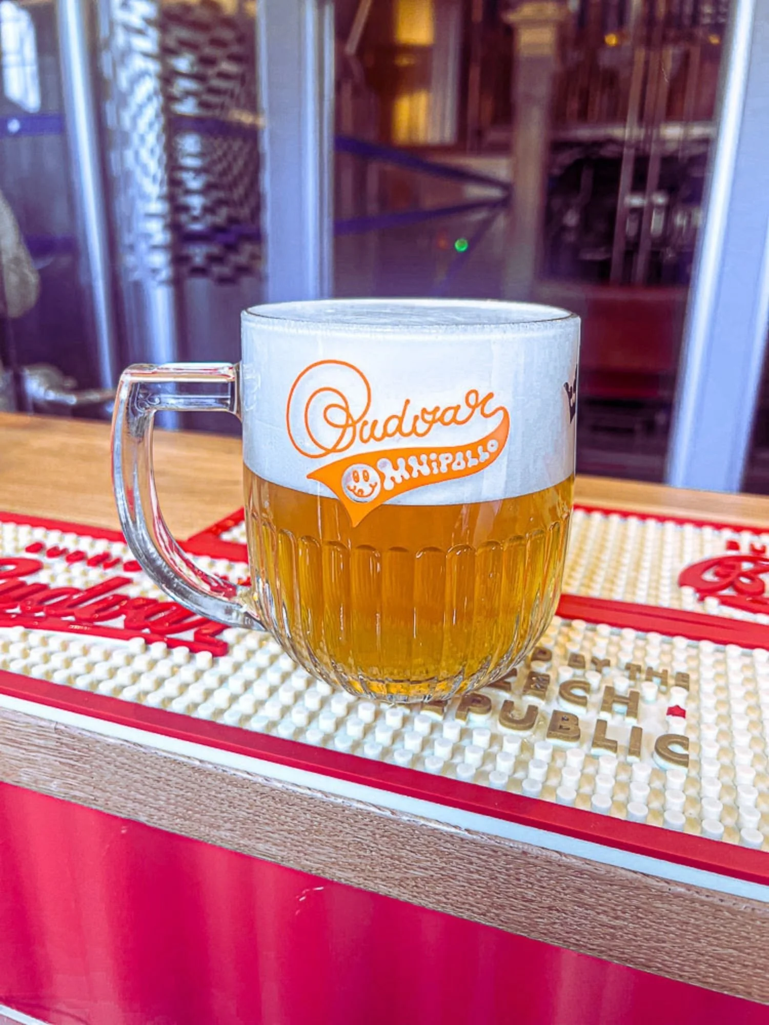 A clear, dimpled glass beer mug filled with golden lager and topped with thick white foam on a bar mat. The glass features an orange co-branded logo reading "Budvar" and "Omnipollo".