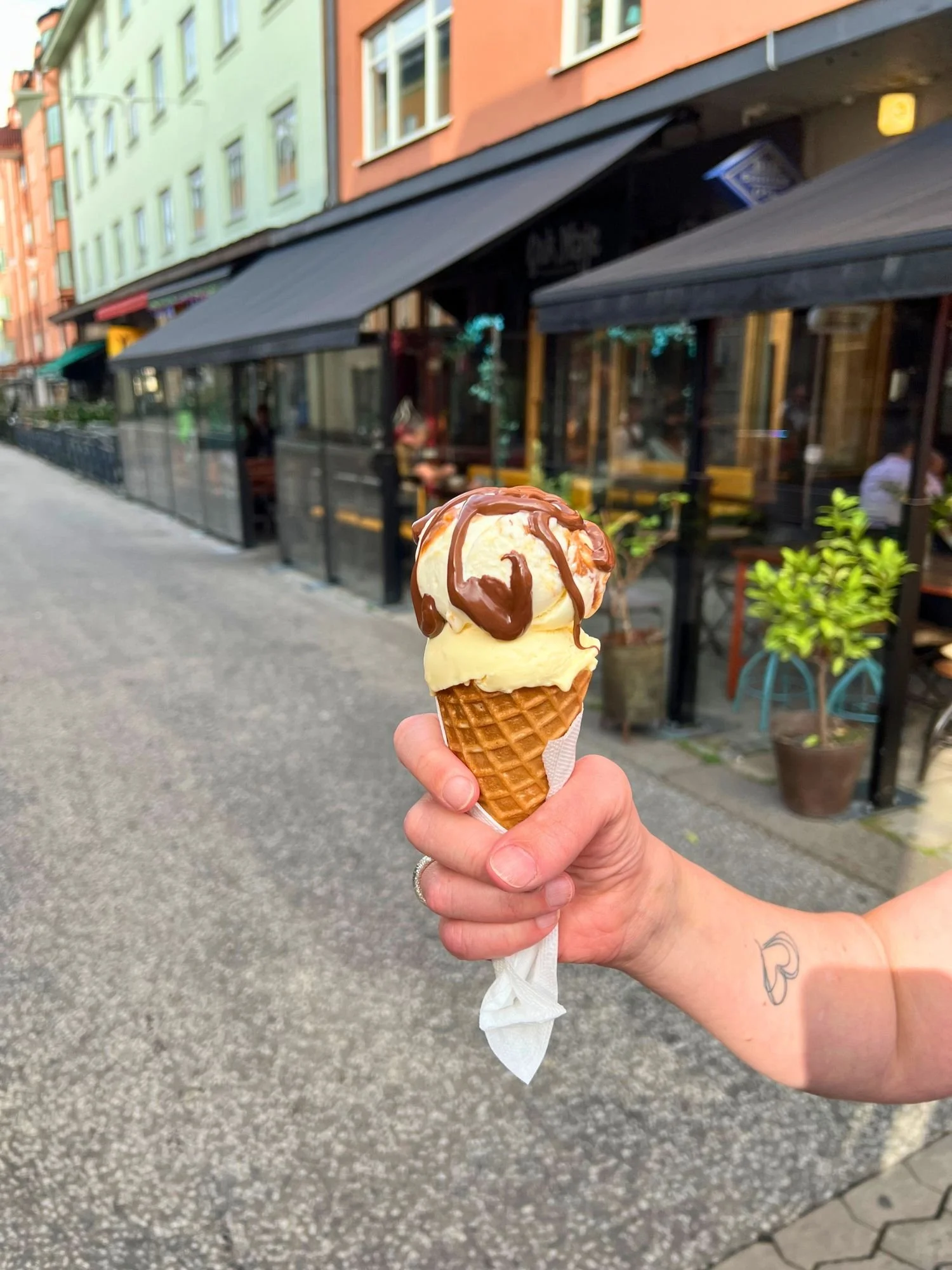 A waffle cone with two scoops of light-colored ice cream and Nutella drizzle, held up against a blurry city street background.