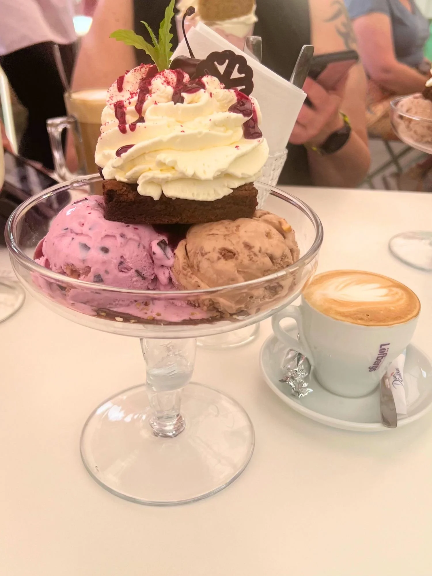 A large ice cream sundae served in a glass dessert bowl with two scoops of ice cream, a brownie, whipped cream, and cherry sauce, alongside a cappuccino. The dessert is served at an outdoor café with people dining in the background.