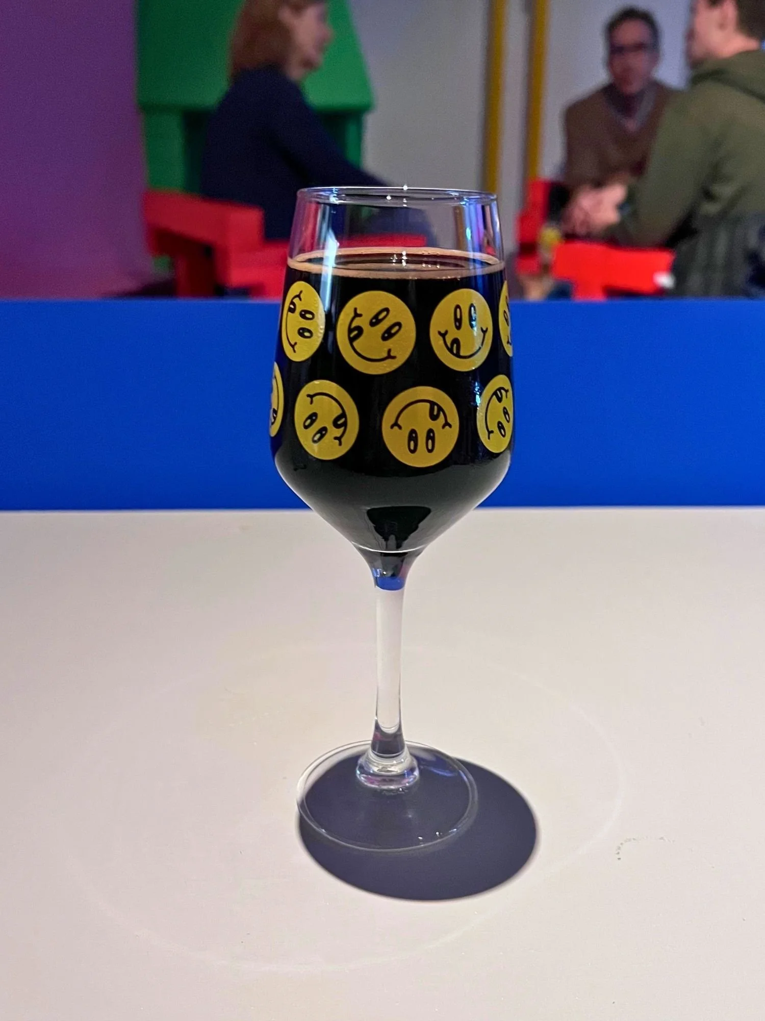 A stemmed glass filled with dark beer sits centered on a smooth white table. The glass is decorated with a pattern of yellow circular smiley faces sticking their tongues out.