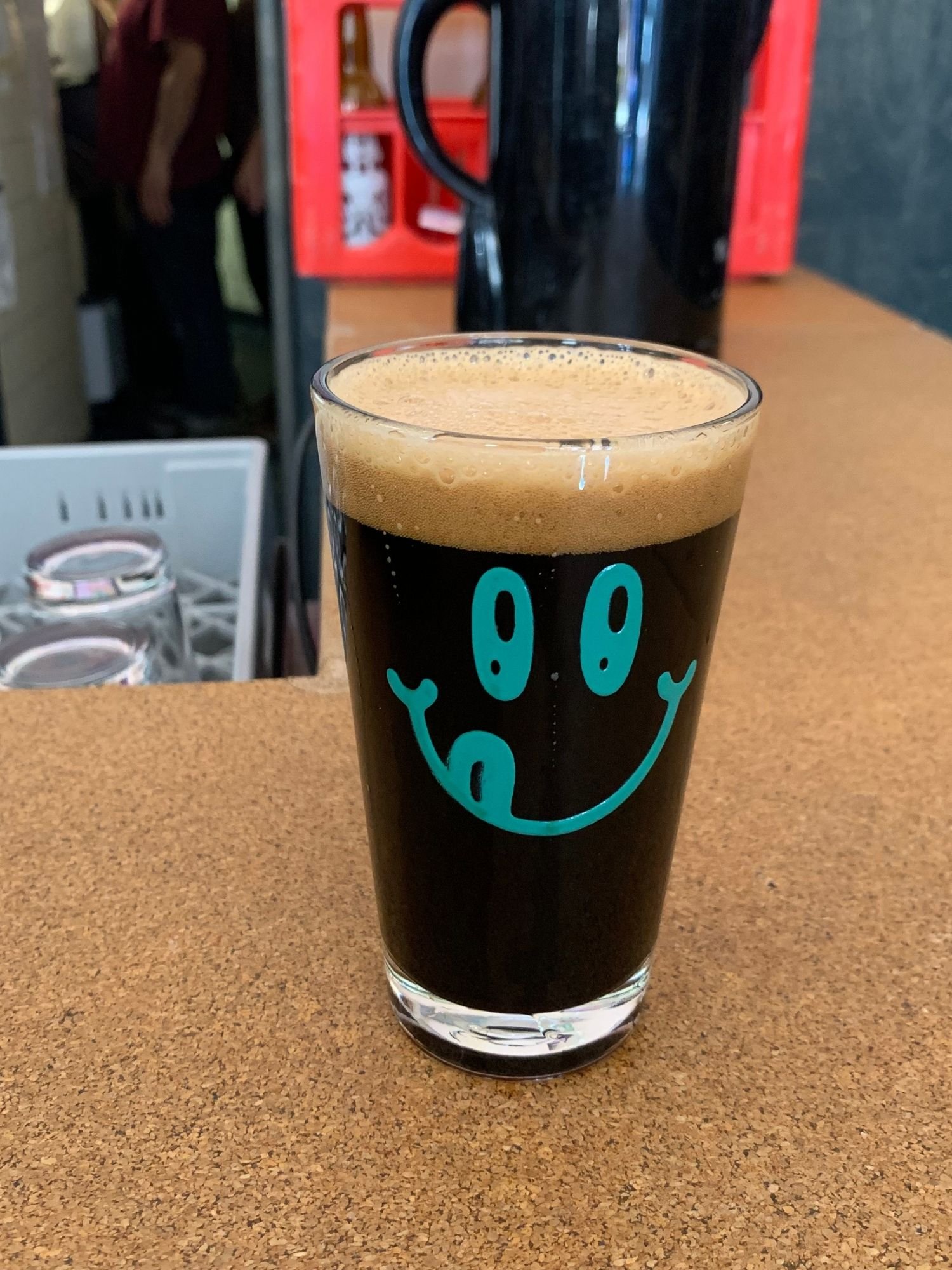 A clear pint glass filled with a dark stout beer topped with a thick, tan foam layer, sitting on a textured brown wooden counter. The glass is printed with a bright teal graphic of a smiley face sticking its tongue out.