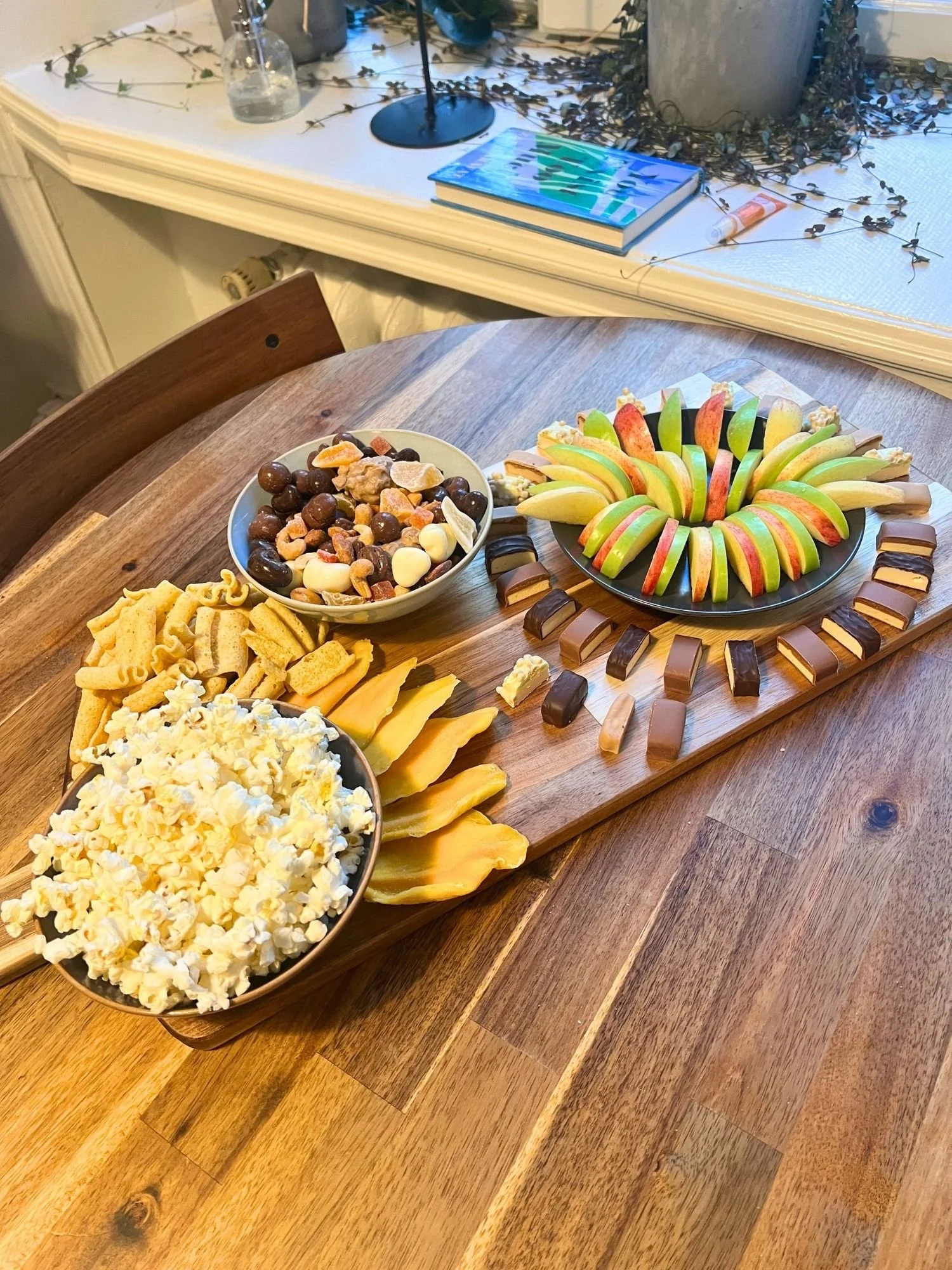 High-angle, length-wise shot of a wooden serving paddle on a round wooden table, laden with popcorn, lentil chips, dried mango, trail mix, sliced apples, and protein bars.