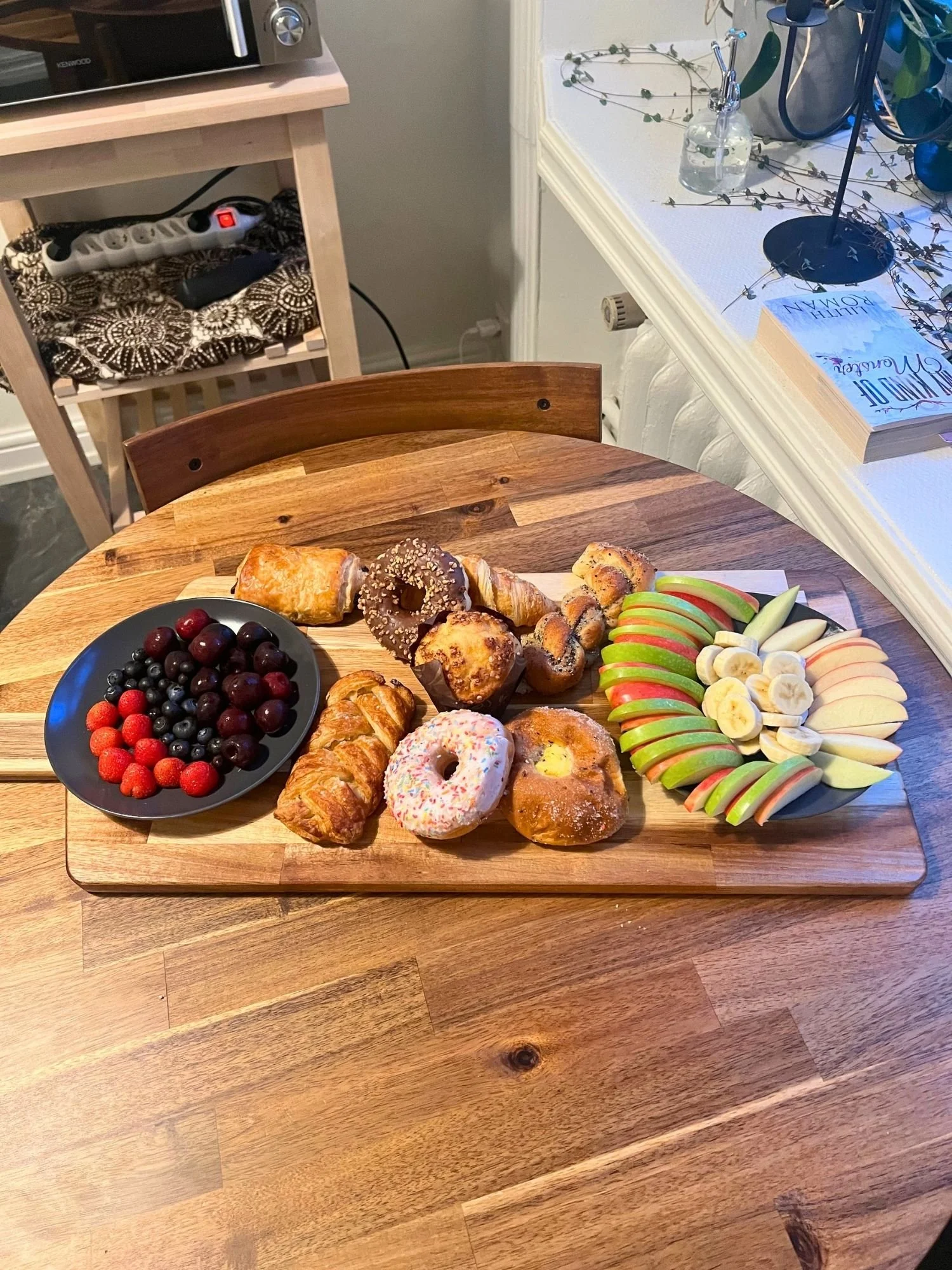 A wooden serving board on a round wooden table filled with an assortment of pastries, donuts, muffins, and plates of fresh fruit.