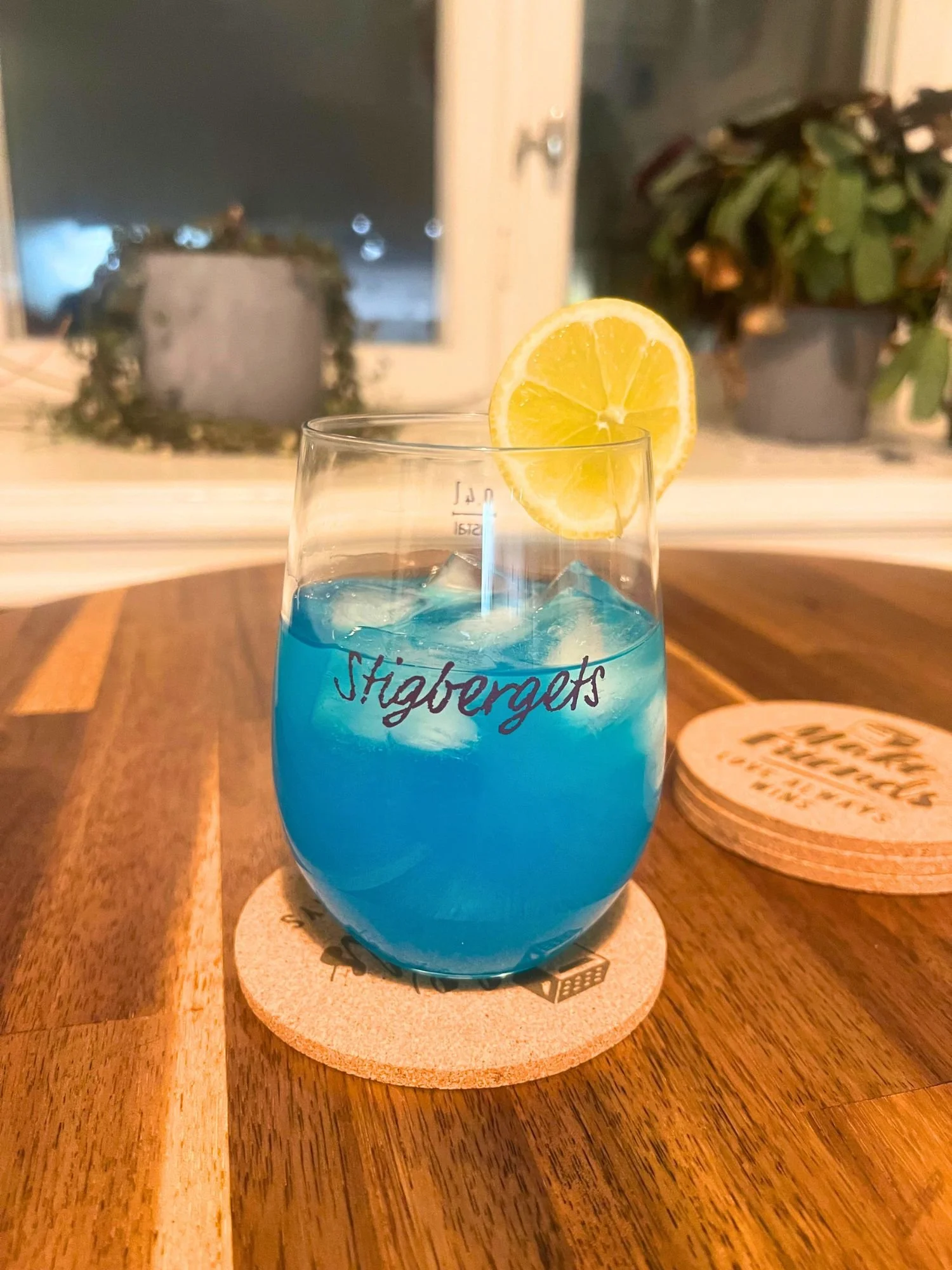Glass of bright blue cocktail over ice, garnished with a lemon slice, placed on a wooden table with a window and plants in the background.