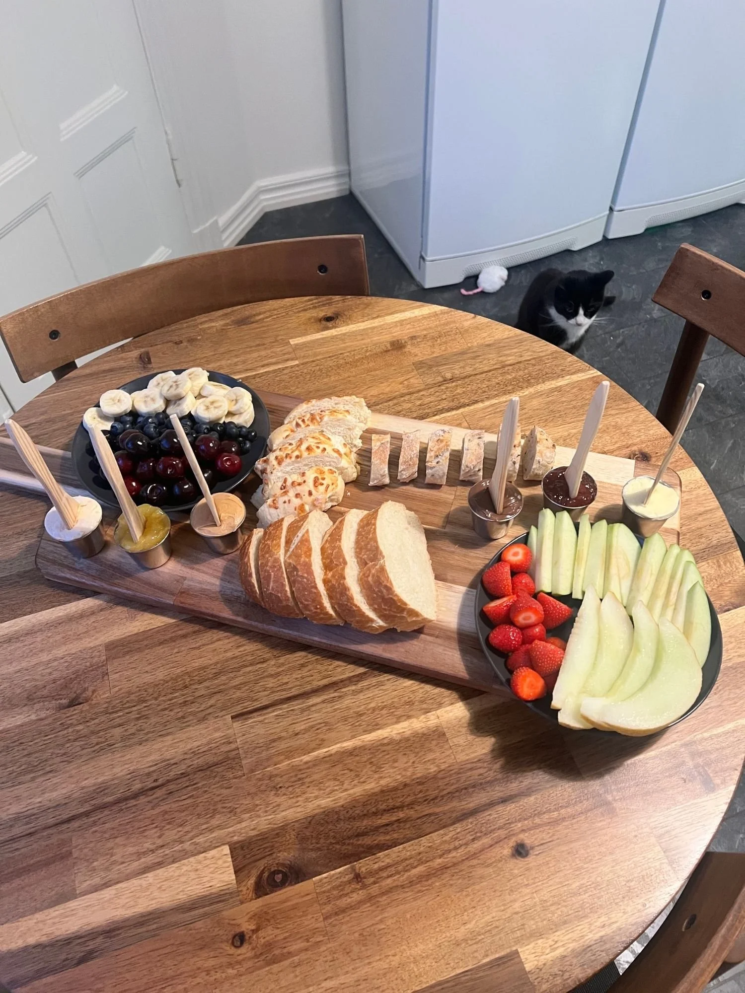 High-angle shot of a wooden board on a round table arranged with sliced bread, fresh fruit, plates with banana, berries, melon, and strawberries, and small cups of spreads with wooden spoons, while a black and white cat sits on the floor nearby.
