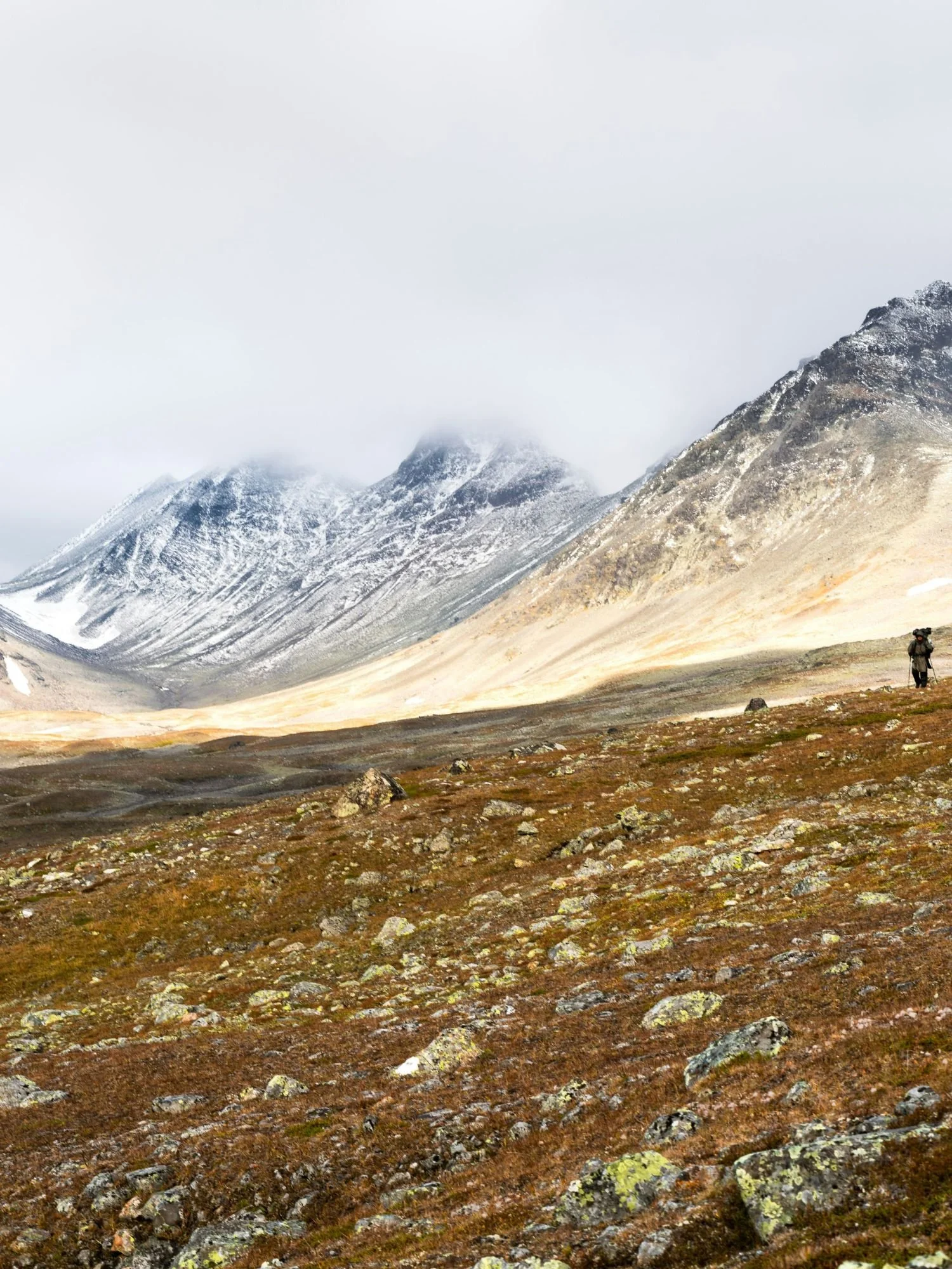 A lone hiker with a large backpack walks across a vast, rocky tundra valley toward massive, snow-capped mountains partially obscured by low clouds in Sarek National Park.