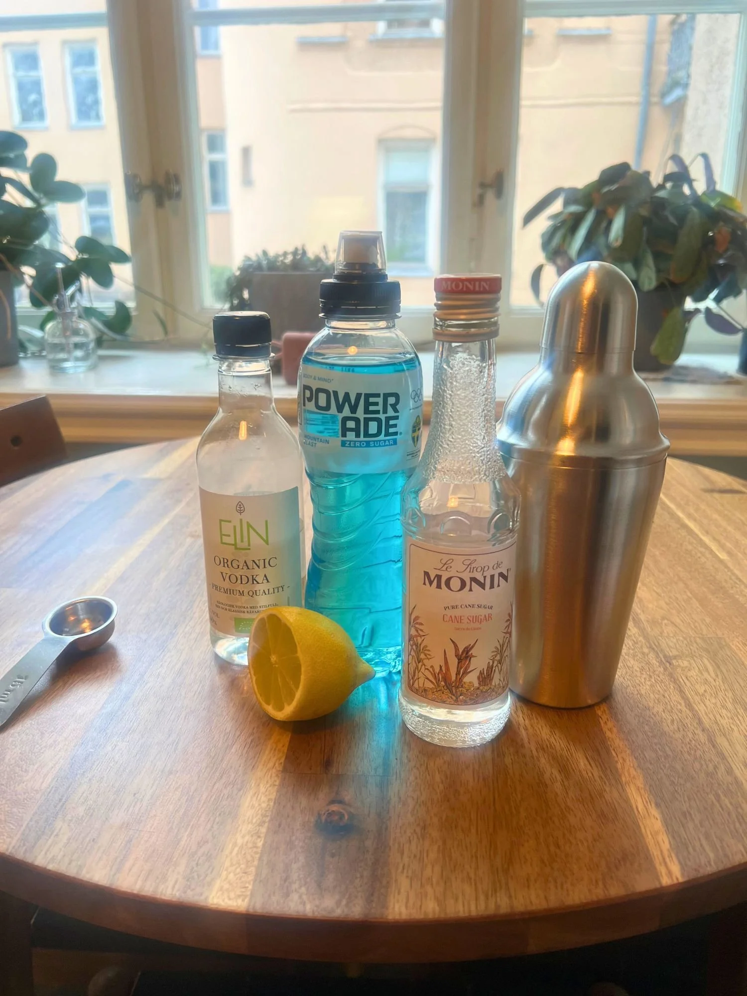 Ingredients for a blue cocktail on a wooden table, including vodka, blue Powerade, simple syrup, a lemon, a cocktail shaker, and a measuring spoon, with a window and plants in the background.