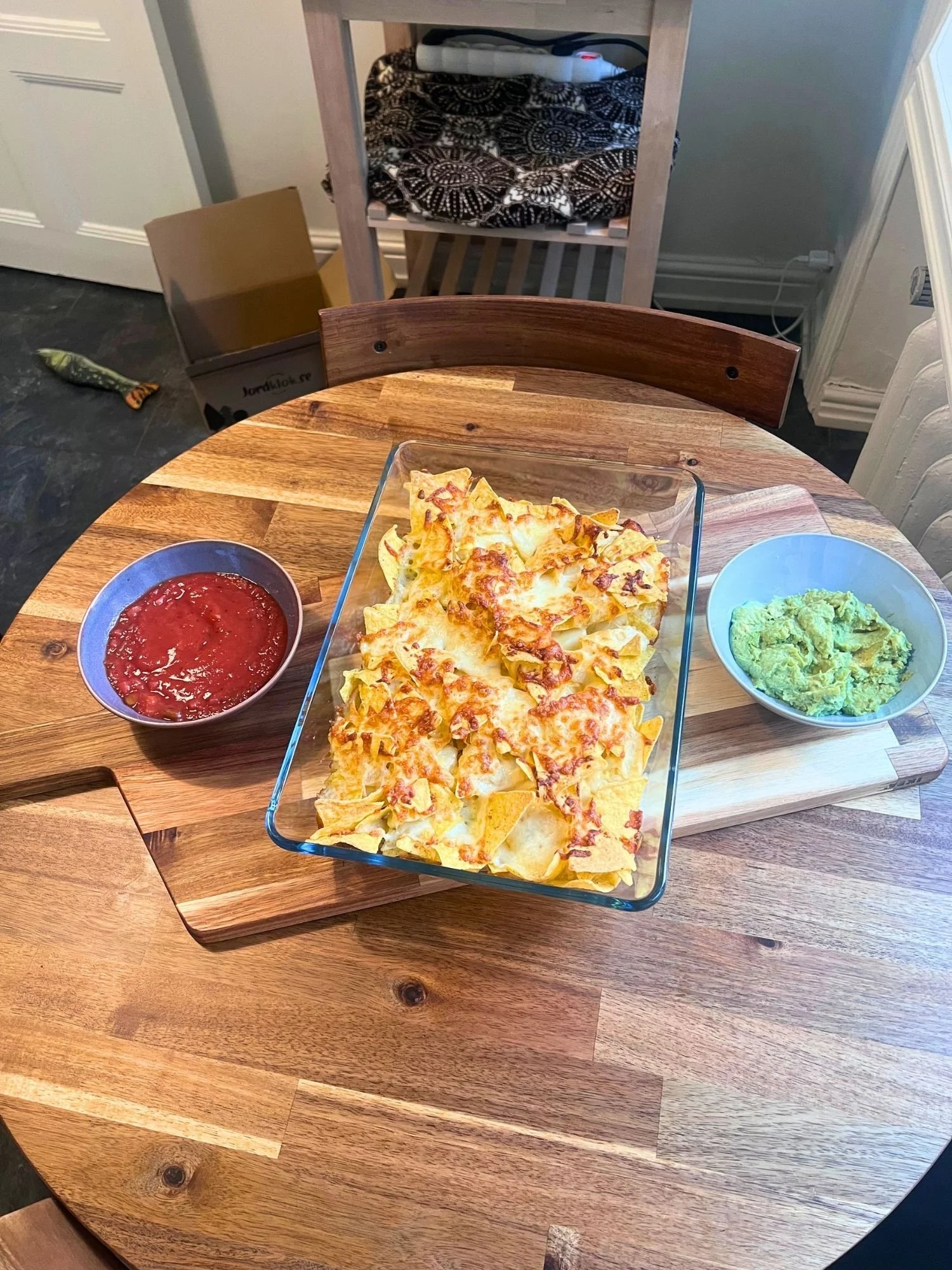 A wooden table set with a glass baking dish of melted cheese nachos, flanked by a bowl of red salsa and a bowl of guacamole on a wooden serving board.