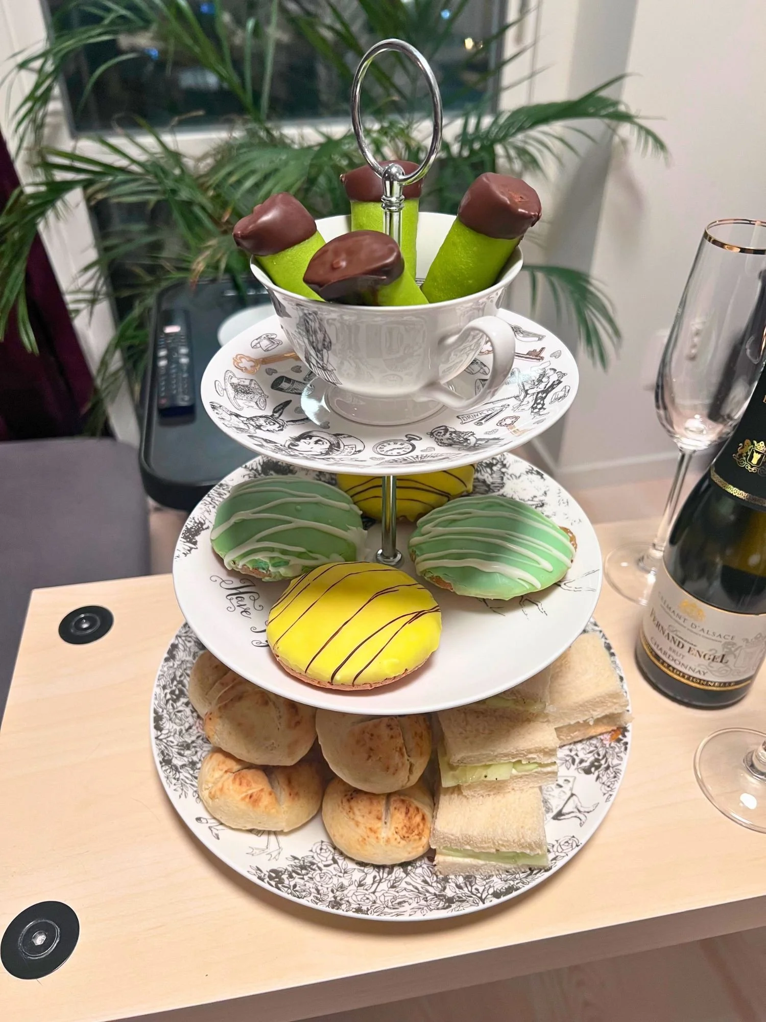 An afternoon tea service on a light wood table. A white teacup with black Alice in Wonderland themed illustrations holds four green dammsugare pastries with chocolate-dipped ends, resting on a matching illustrated saucer.