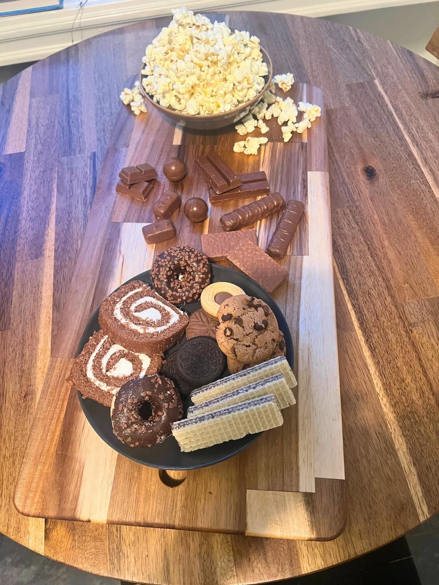 High-angle, top-down shot of a wooden serving board set on a round wooden table, holding a plate of assorted cookies and mini donuts, loose chocolates, and a bowl of popcorn.