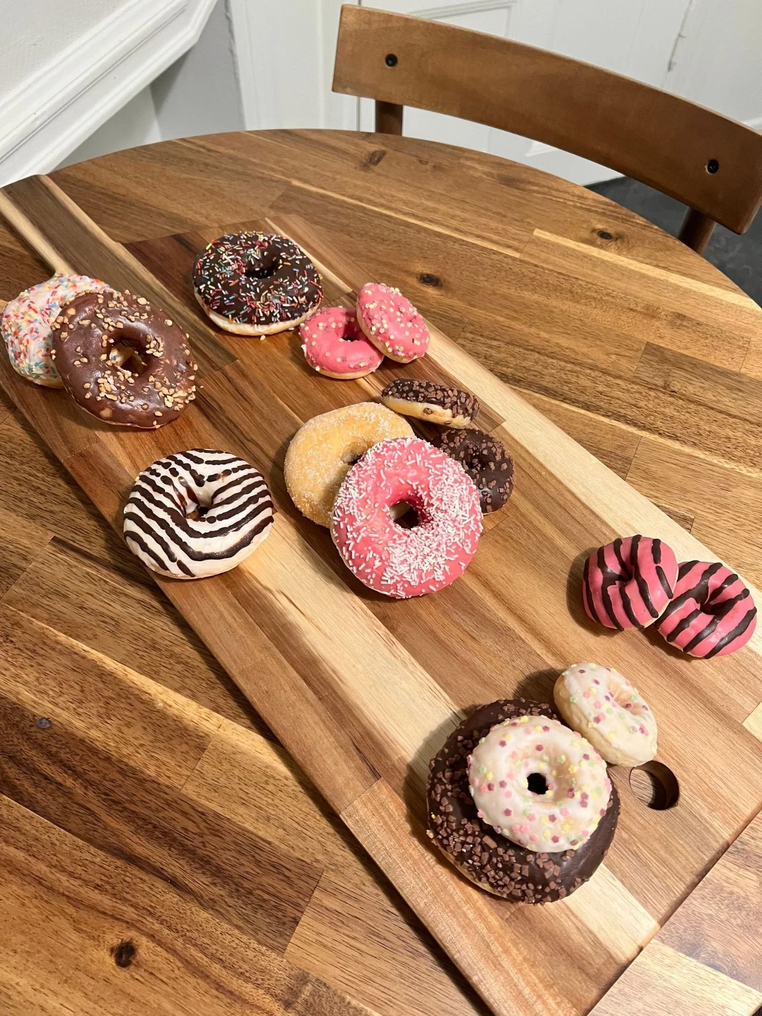 A long wooden serving board rests diagonally across a round wooden table, colorful and varied donuts scattered across its surface.