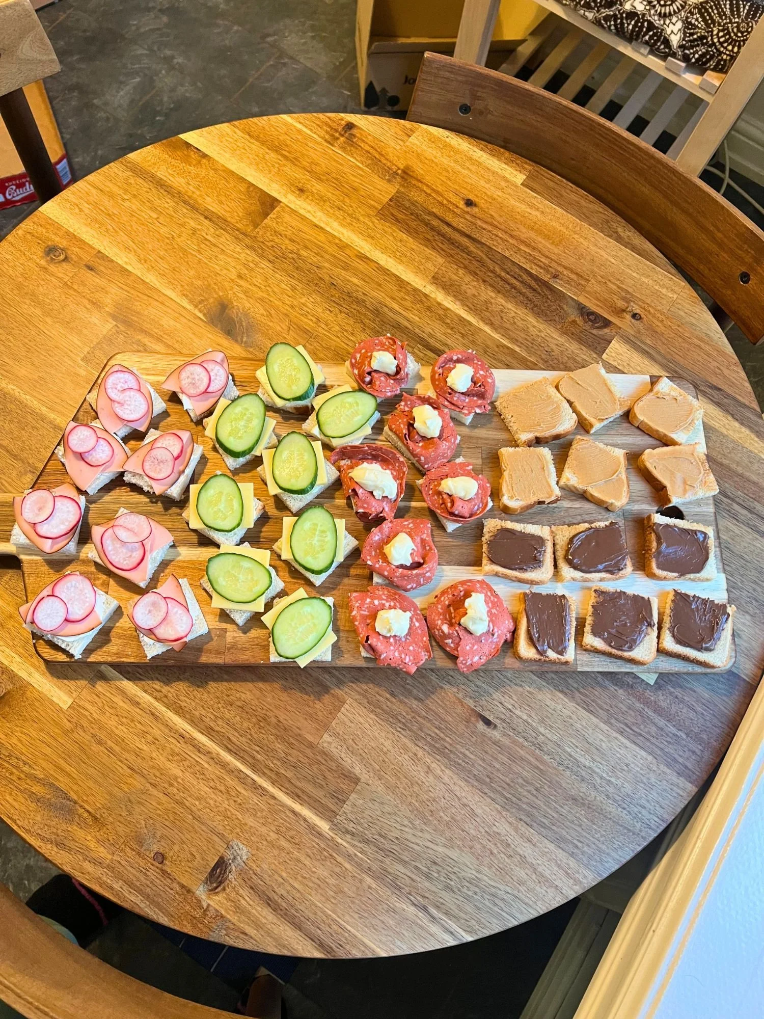 High-angle shot of a rectangular wooden serving board on a round wooden table, neatly arranged with columns of small, bite-sized open-face sandwiches.