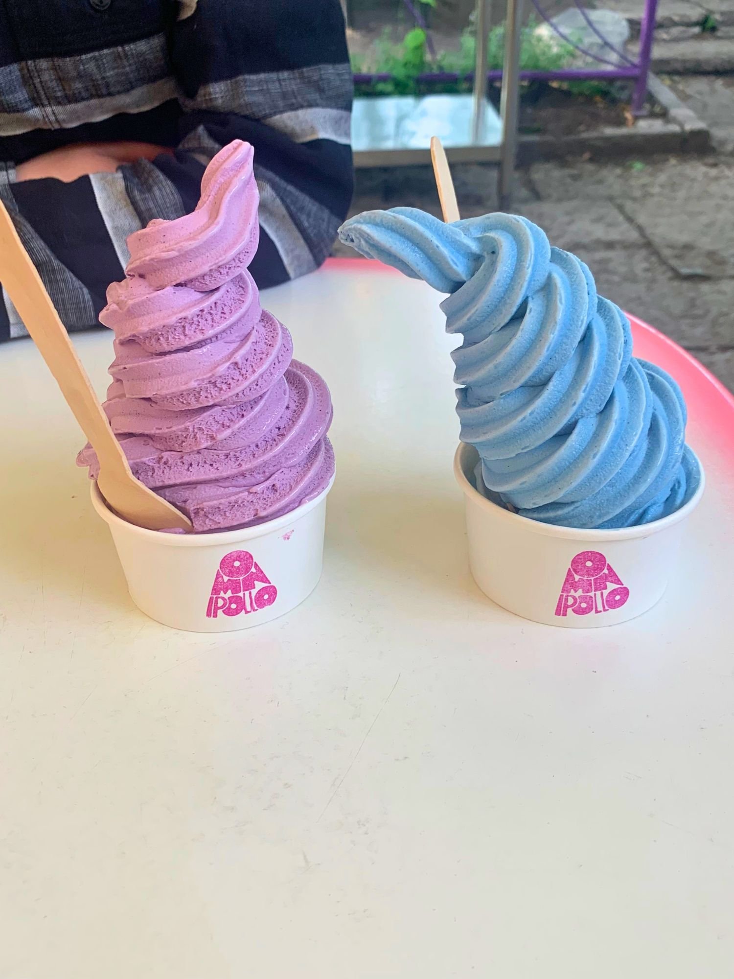 Two vibrant cups of soft-serve ice cream side-by-side on a white table. The cup on the left has a tall swirl of purple ice cream and a wooden spoon. The cup on the right has a tall, leaning swirl of bright blue ice cream.