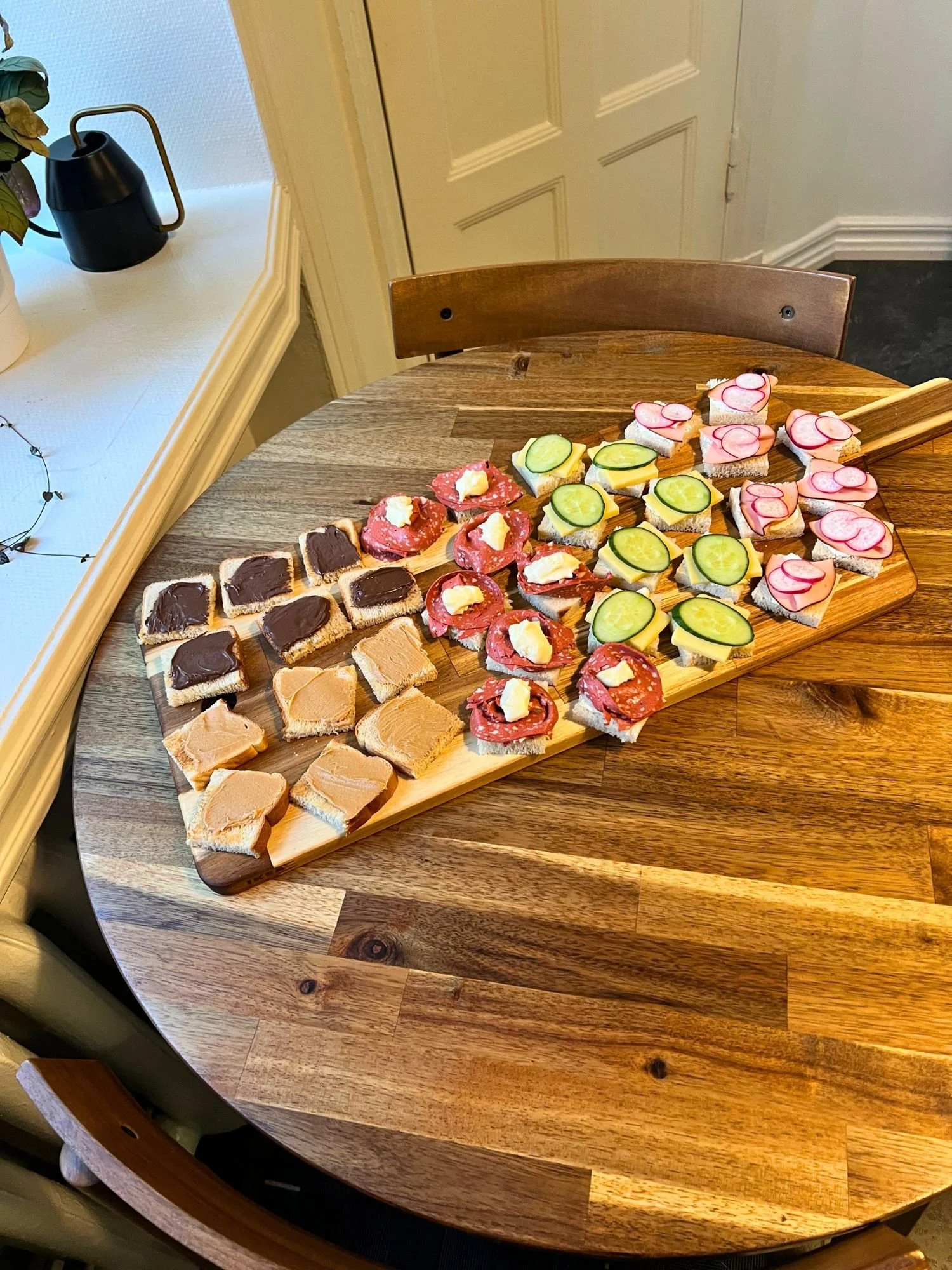 High-angle shot of a long wooden serving board on a round wooden table, neatly arranged with rows of bite-sized open-face mini sandwiches with various toppings.