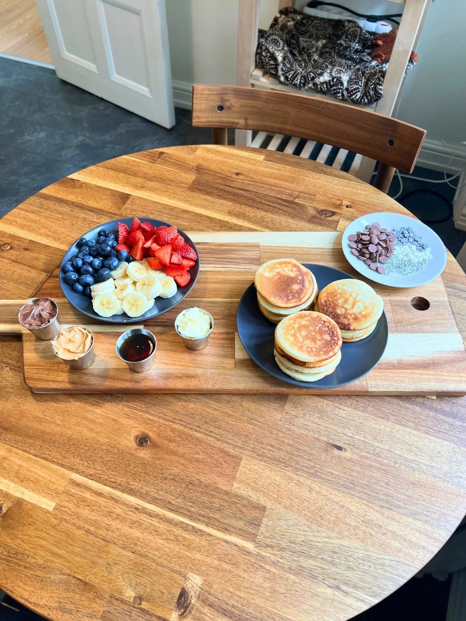 A wooden serving board on a wooden table arranged with stacks of mini pancakes, fresh fruit, toppings, and small cups of spreads and syrup.