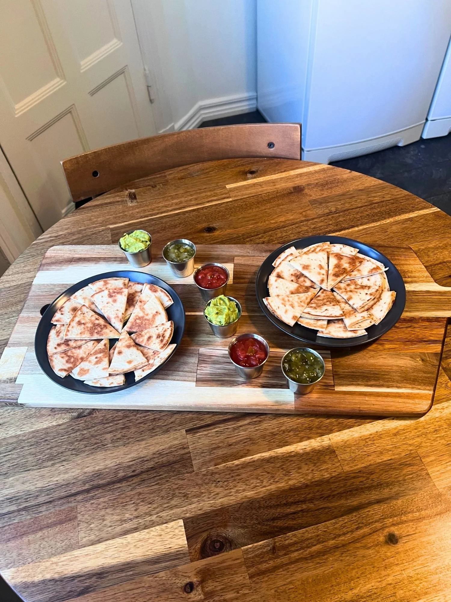 High-angle shot of a rectangular wooden serving board on a round wooden table, set with two dark plates of cut quesadilla wedges and small metal cups of salsa and guacamole.