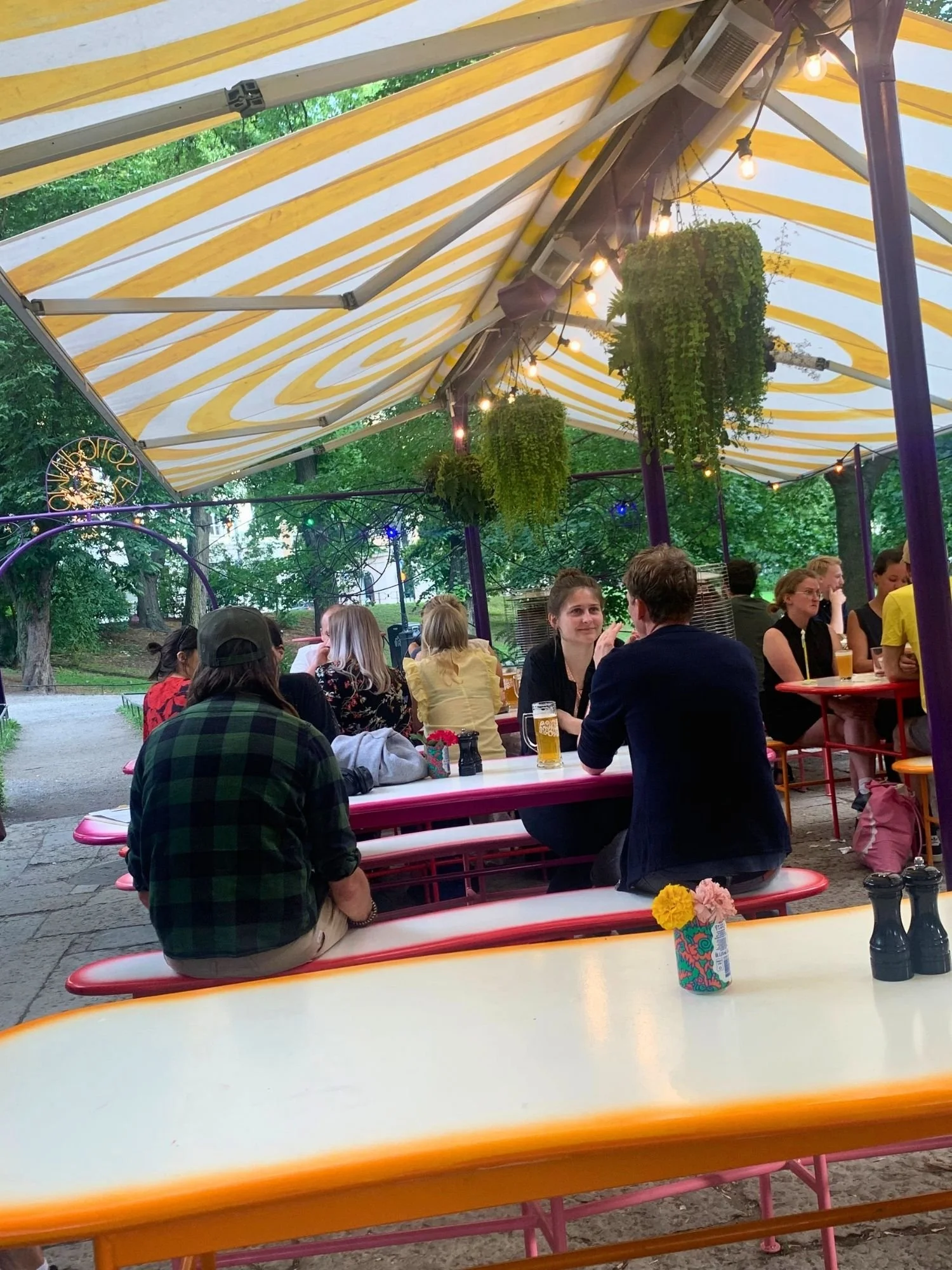 People sit at long outdoor picnic tables under a yellow-and-white striped canopy adorned with hanging string lights and potted plants. In the foreground, a person in a green-and-black plaid shirt sits with their back to the camera at a table.