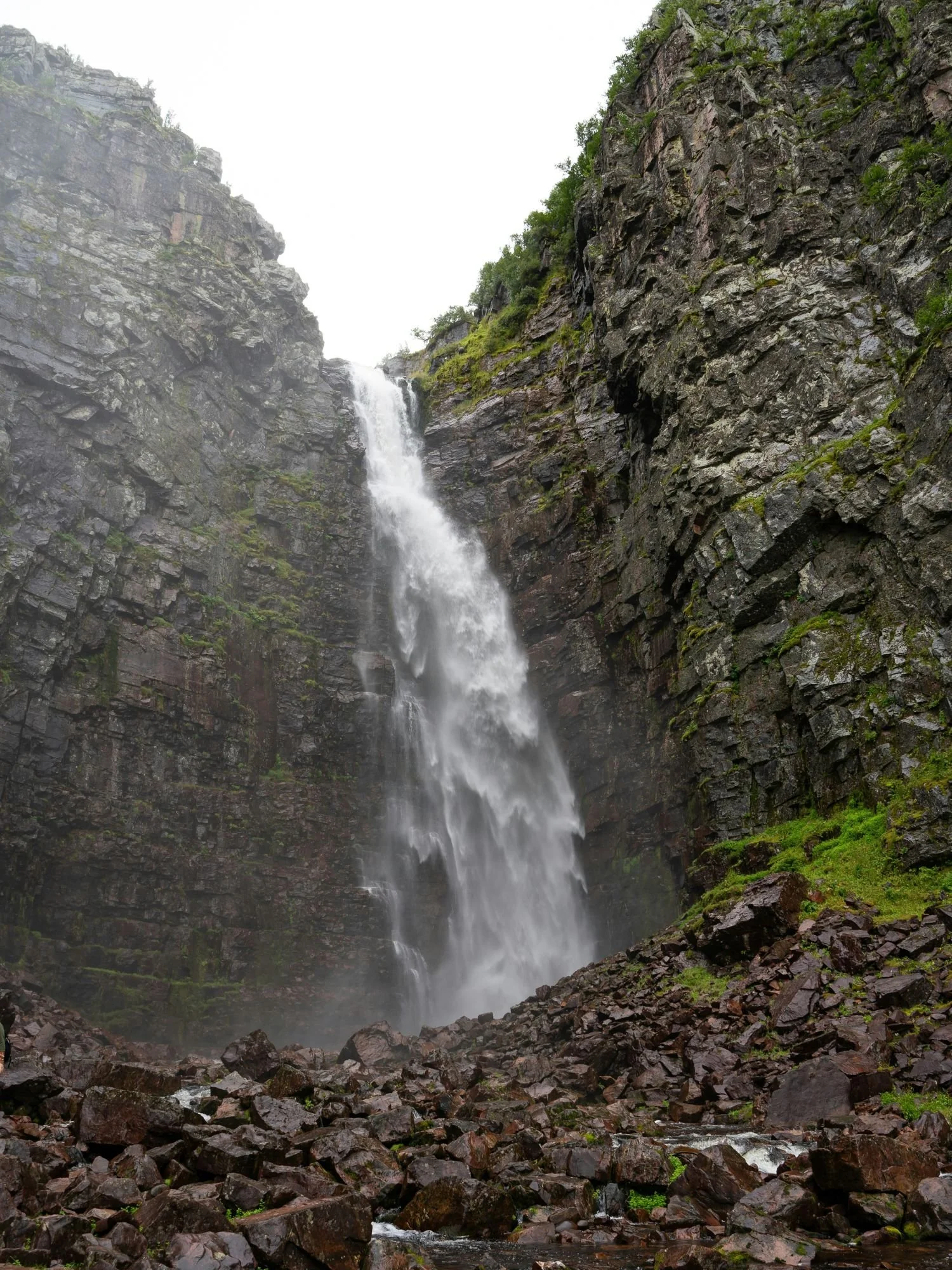 A tall waterfall cascading down a steep rocky cliff surrounded by moss and greenery, with mist rising over scattered stones below.