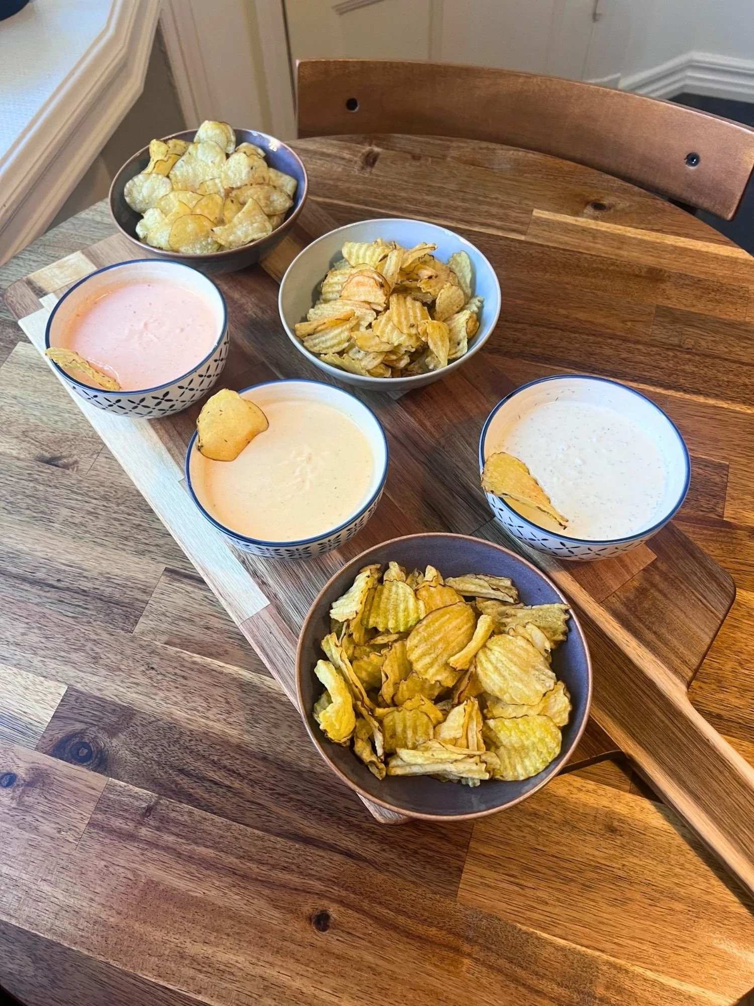 High-angle shot of a wooden serving board set on a round wooden table, holding three bowls of potato chips and three ceramic bowls of creamy dips.