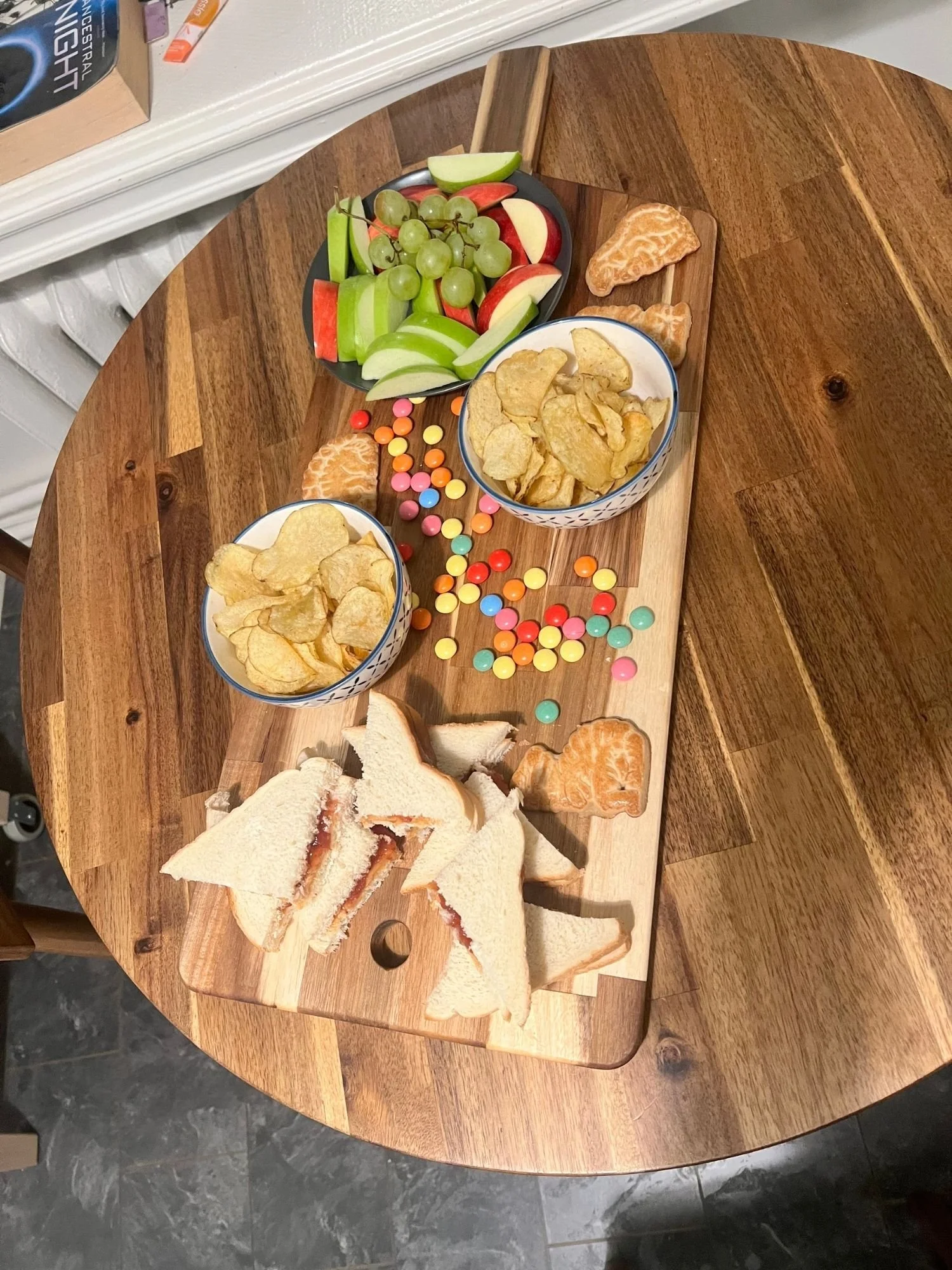 A long wooden serving board lies vertically across a round wooden table, presenting a colorful, kid-friendly snack spread.