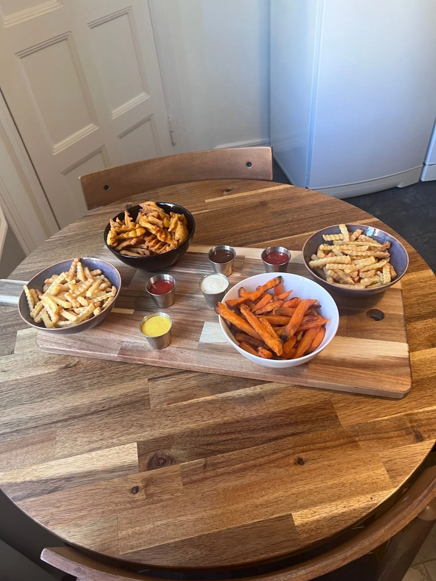 A wooden table set with a serving board holding four bowls of different style fries and sweet potato fries, alongside five small metal ramekins of dipping sauces.