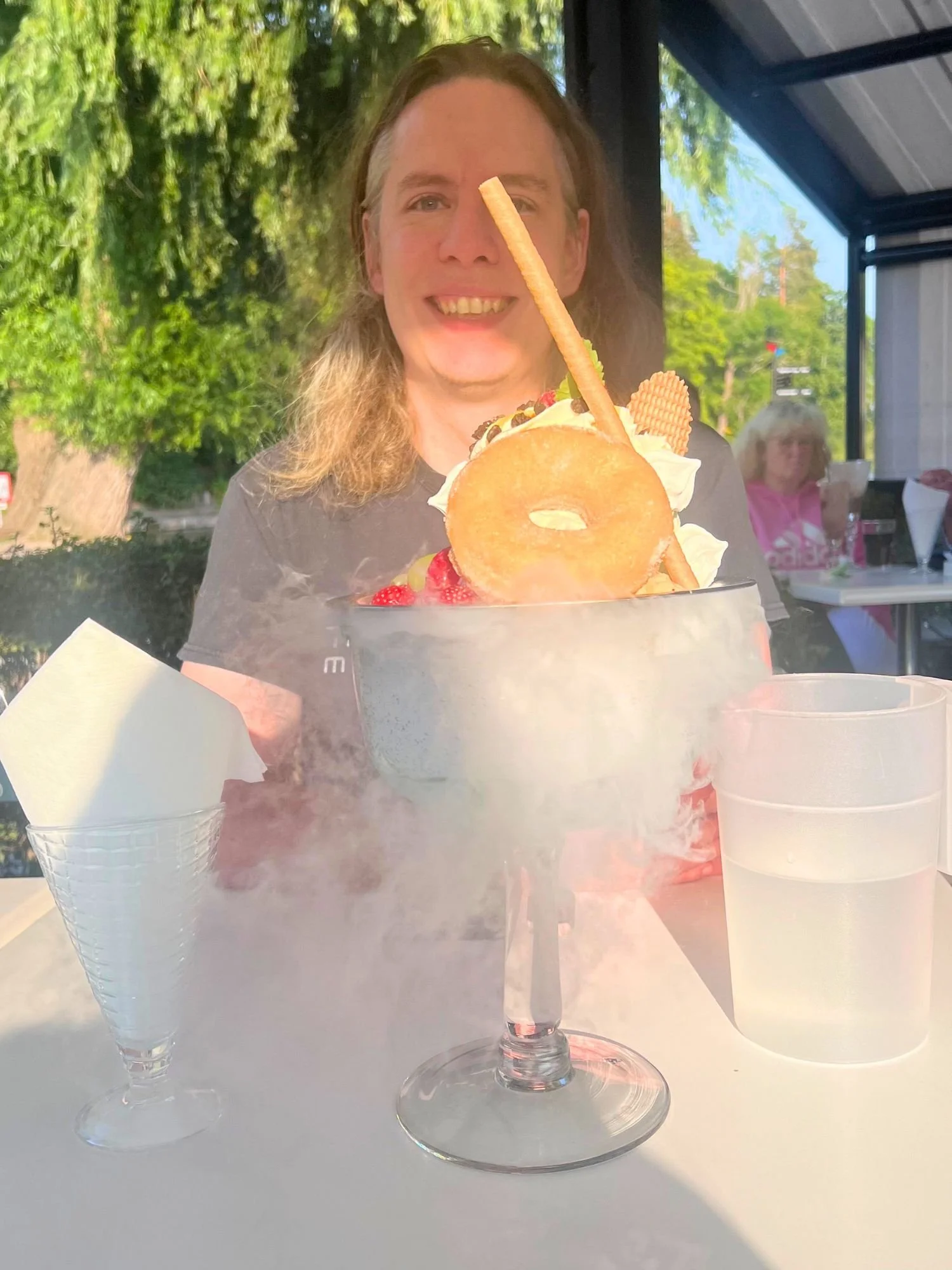 A man with long blonde-brown hair smiling behind an elaborate, oversized ice cream sundae served in a large stemmed goblet.