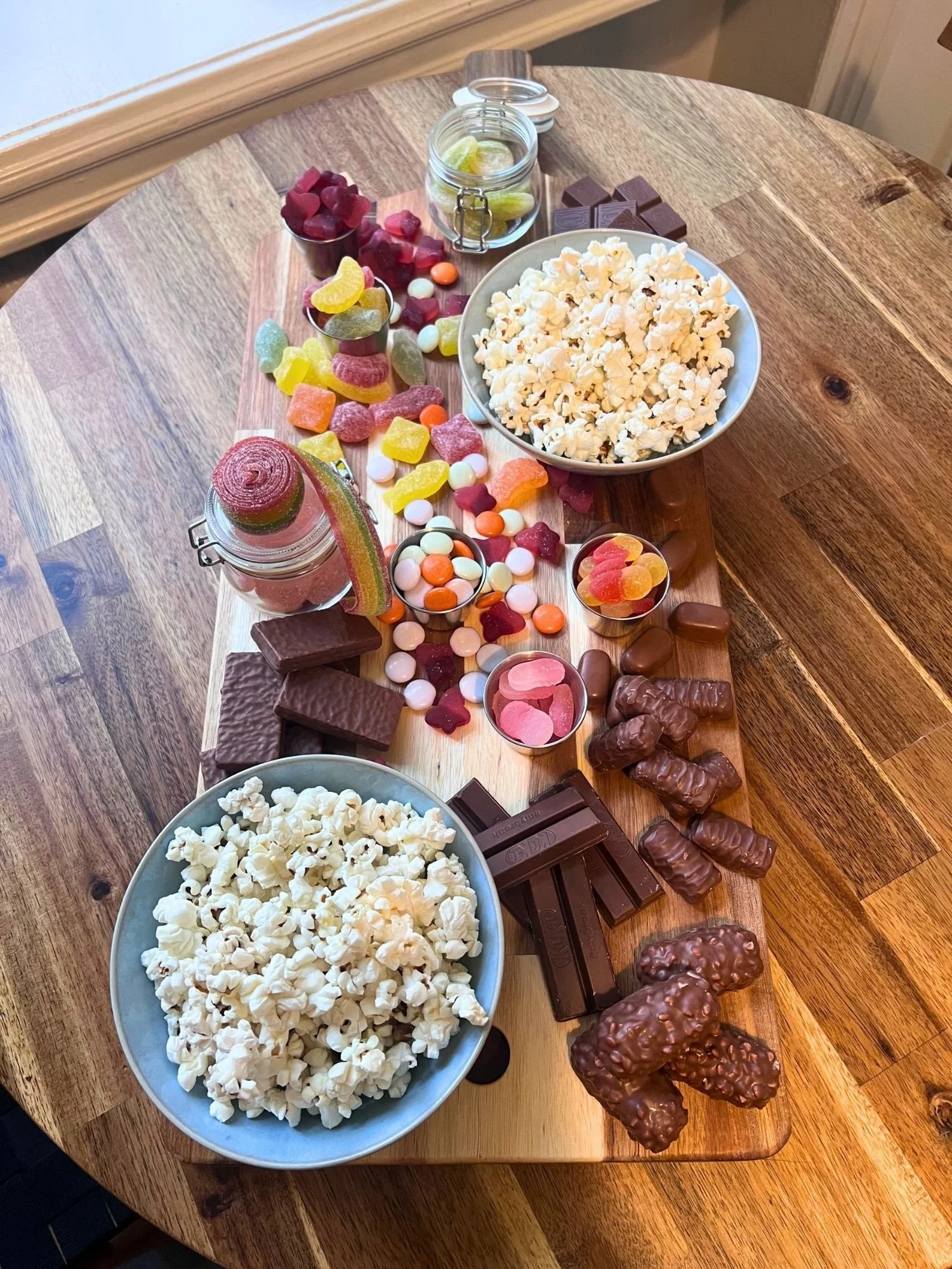wooden table set with a snack board featuring two bowls of popcorn, glass jars of gummies, chocolate bars, and candy-coated chocolates.