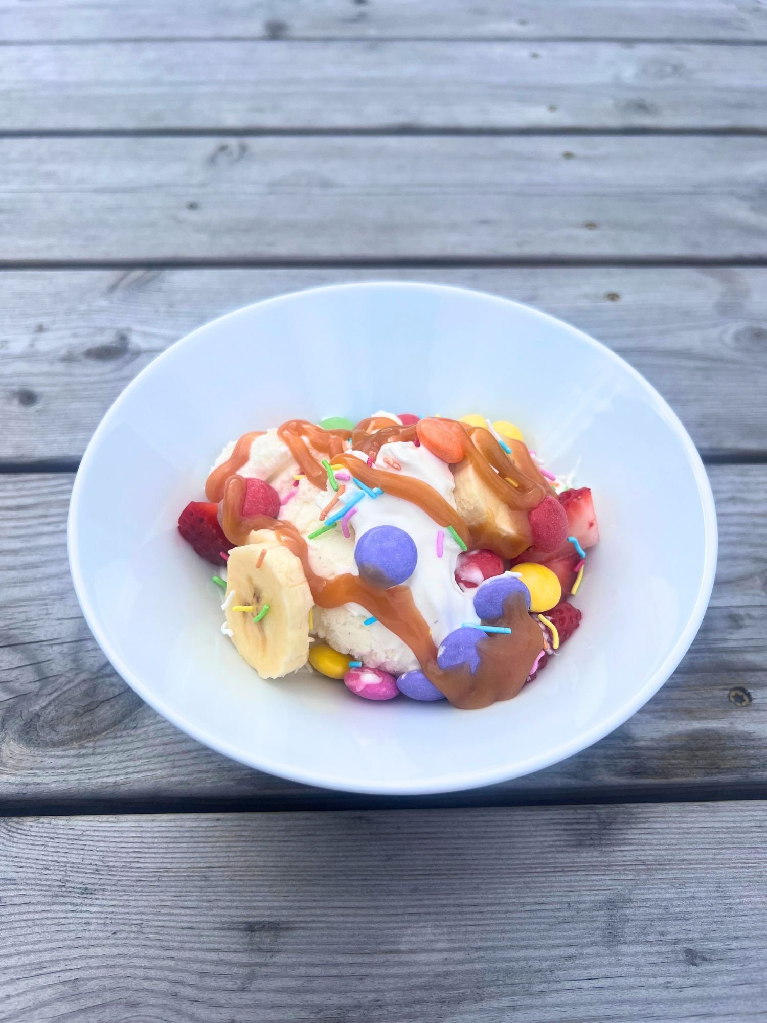 A white bowl filled with vanilla ice cream on a grey wooden table. The ice cream is topped with caramel sauce, banana slices, strawberry pieces, rainbow sprinkles, and colorful candy-coated chocolates.