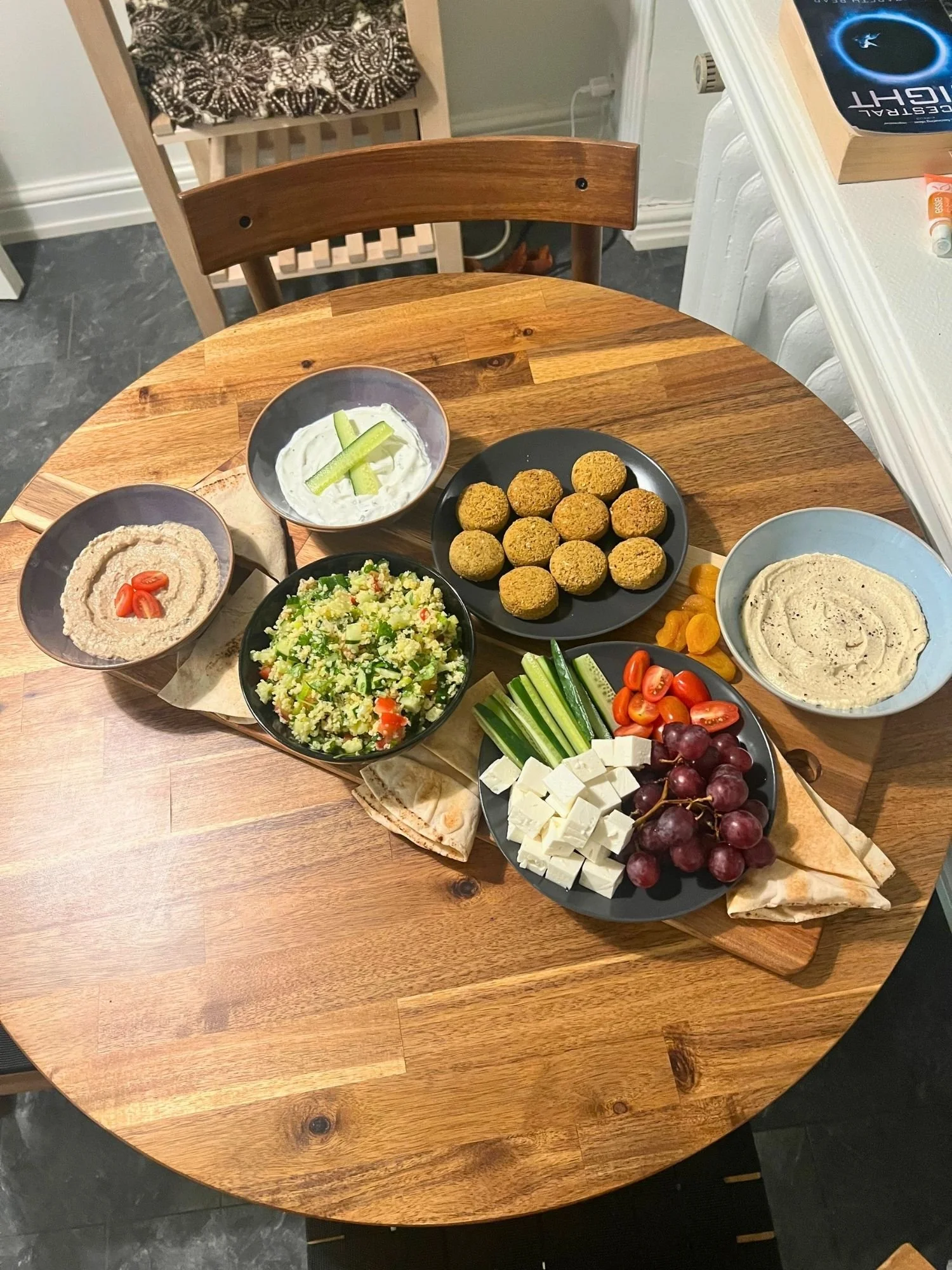 A wooden board rests diagonally across a round wooden table, laden with a Mediterranean-style grazing platter.