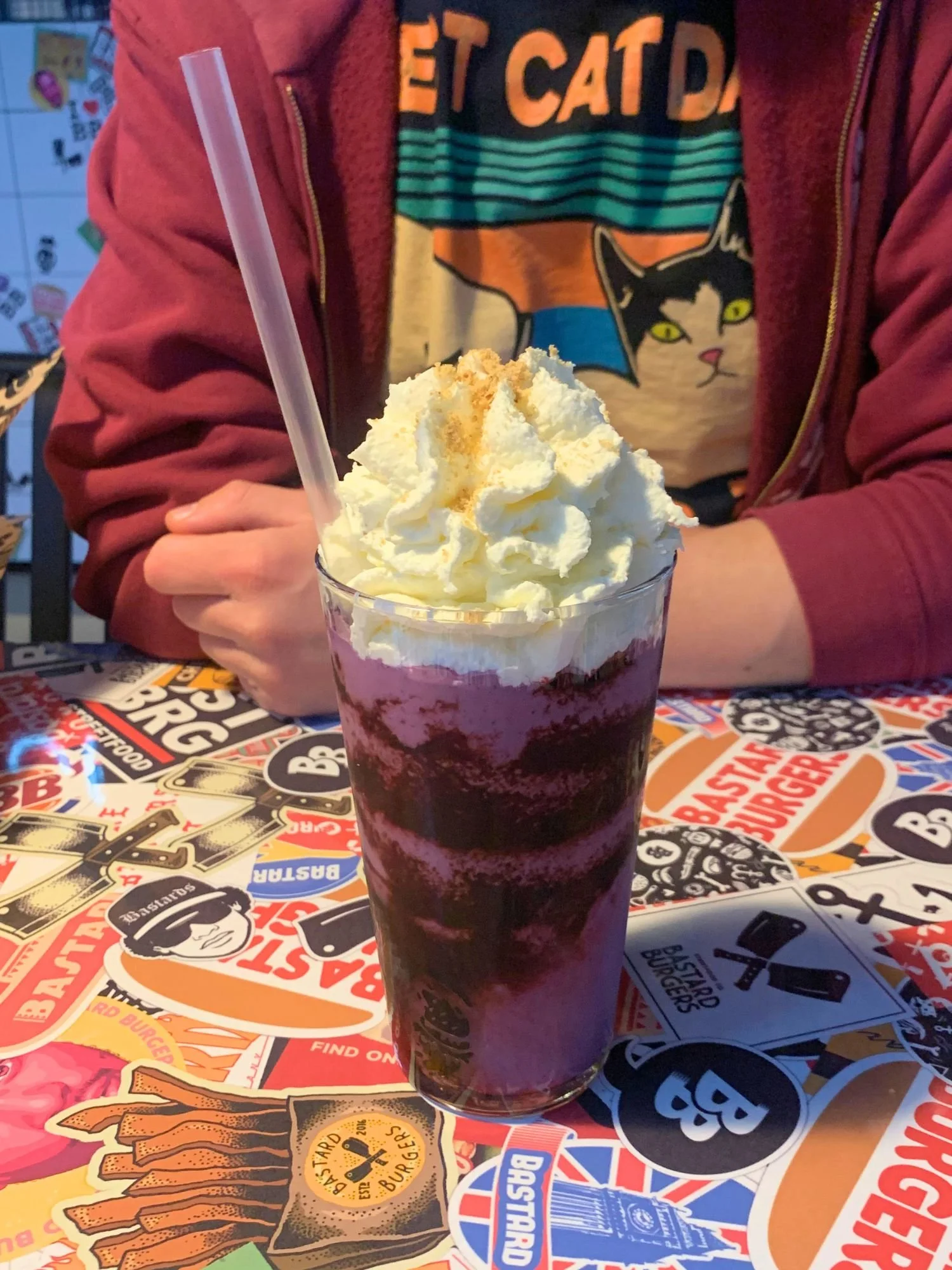 A tall glass filled with a layered purple blueberry milkshake, heavily topped with whipped cream and graham cracker crumbs. A clear straw is inserted into the shake.