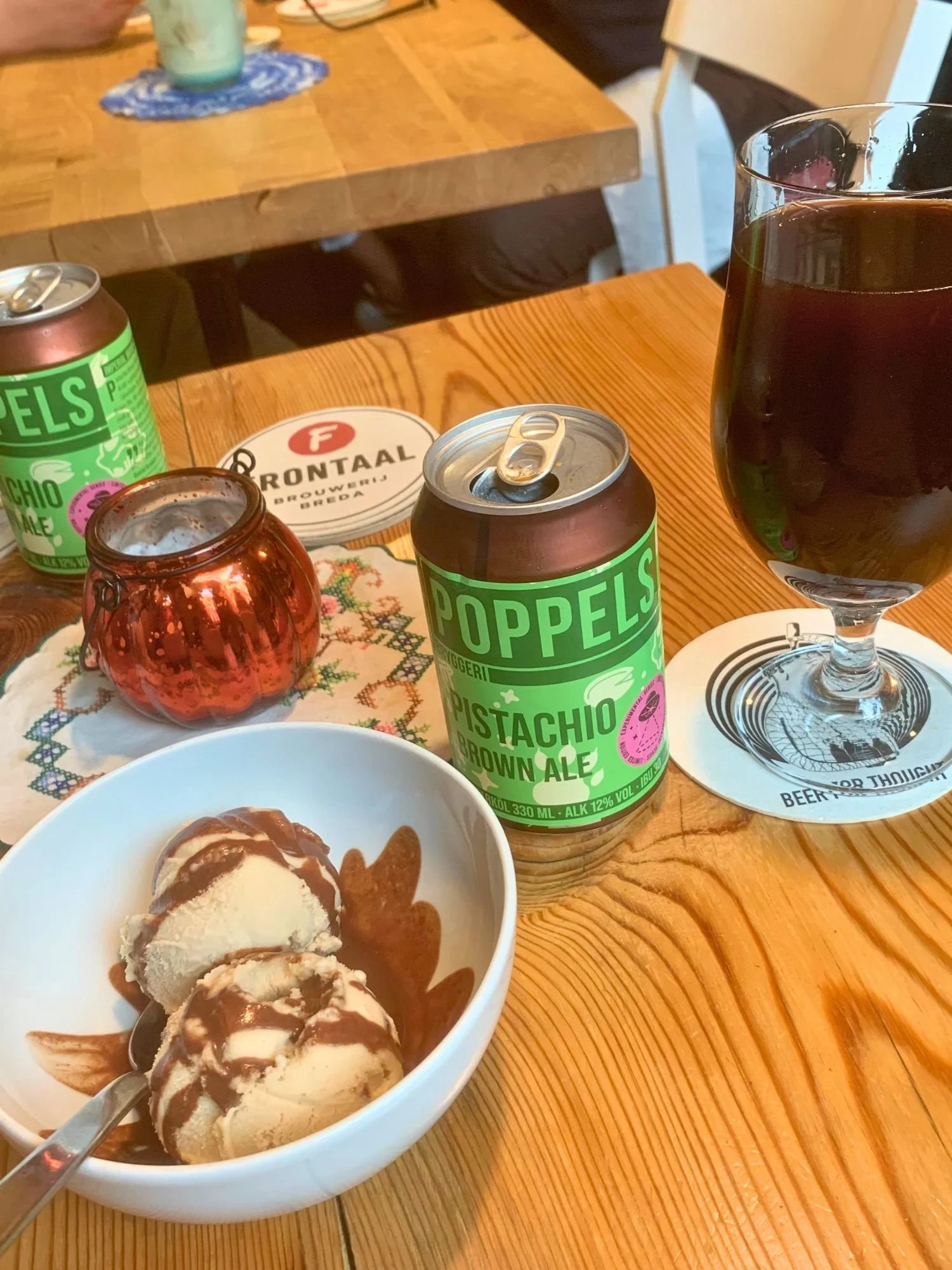 Two scoops of ice cream with chocolate sauce served next to a can and glass of Poppels Pistachio Brown Ale on a wooden table.