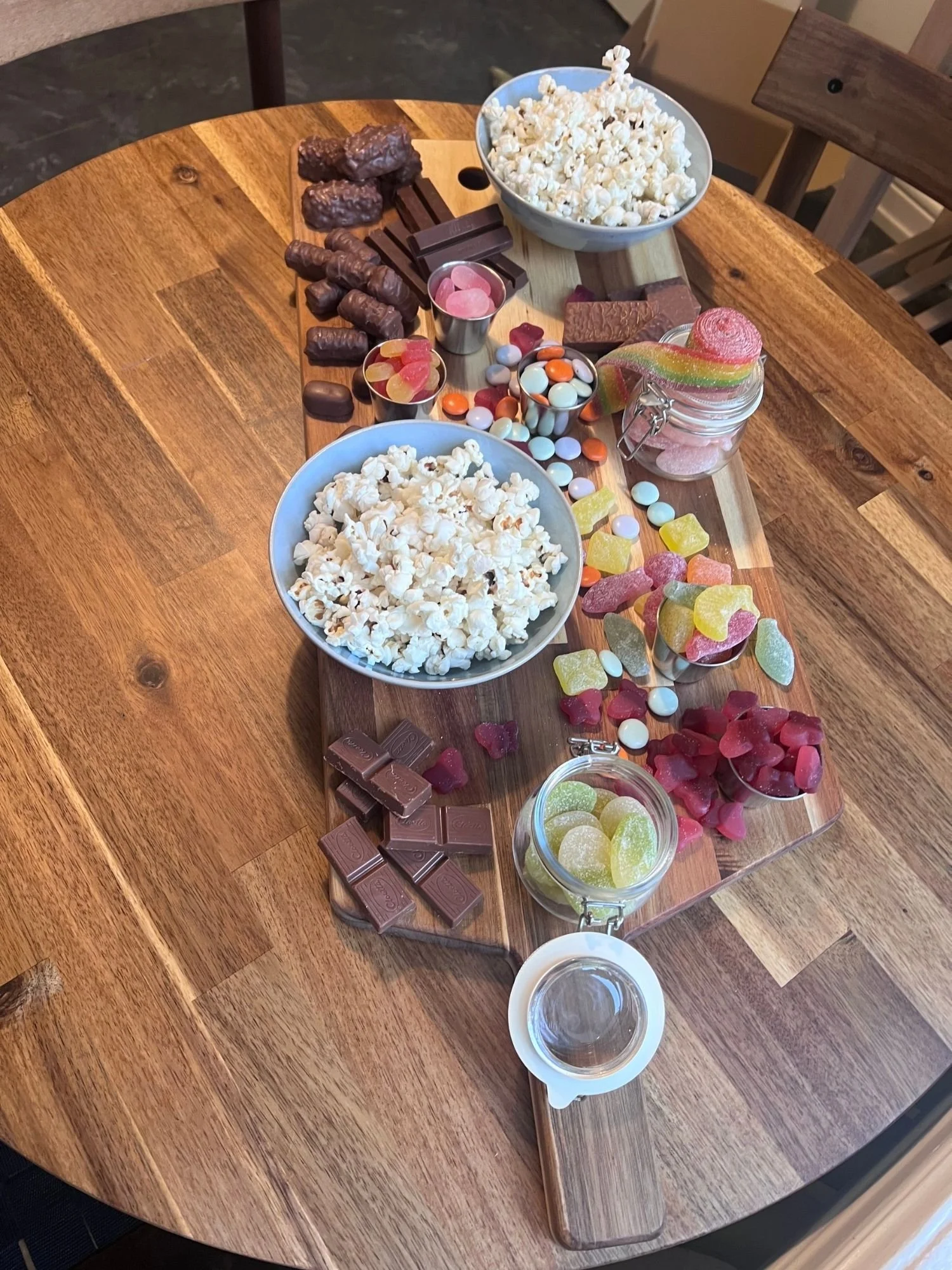 A wooden table set with a long snack board filled with two bowls of popcorn, glass jars of gummies, chocolate bars, and candy-coated chocolates.
