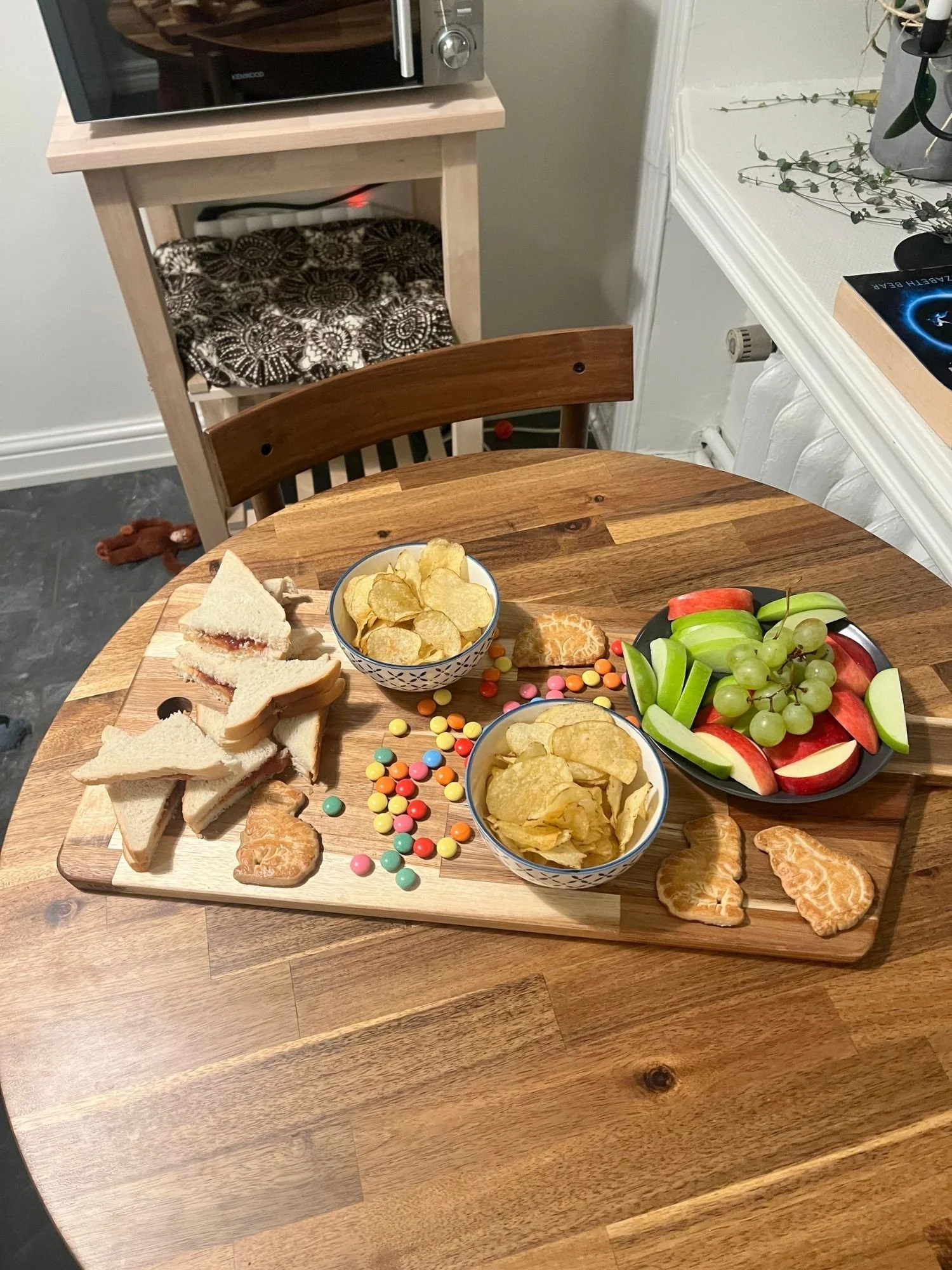A kid-friendly lunch platter on a wooden board featuring sandwich triangles, two bowls of potato chips, dinosaur cookies, colorful candy, and a plate of sliced apples and green grapes.