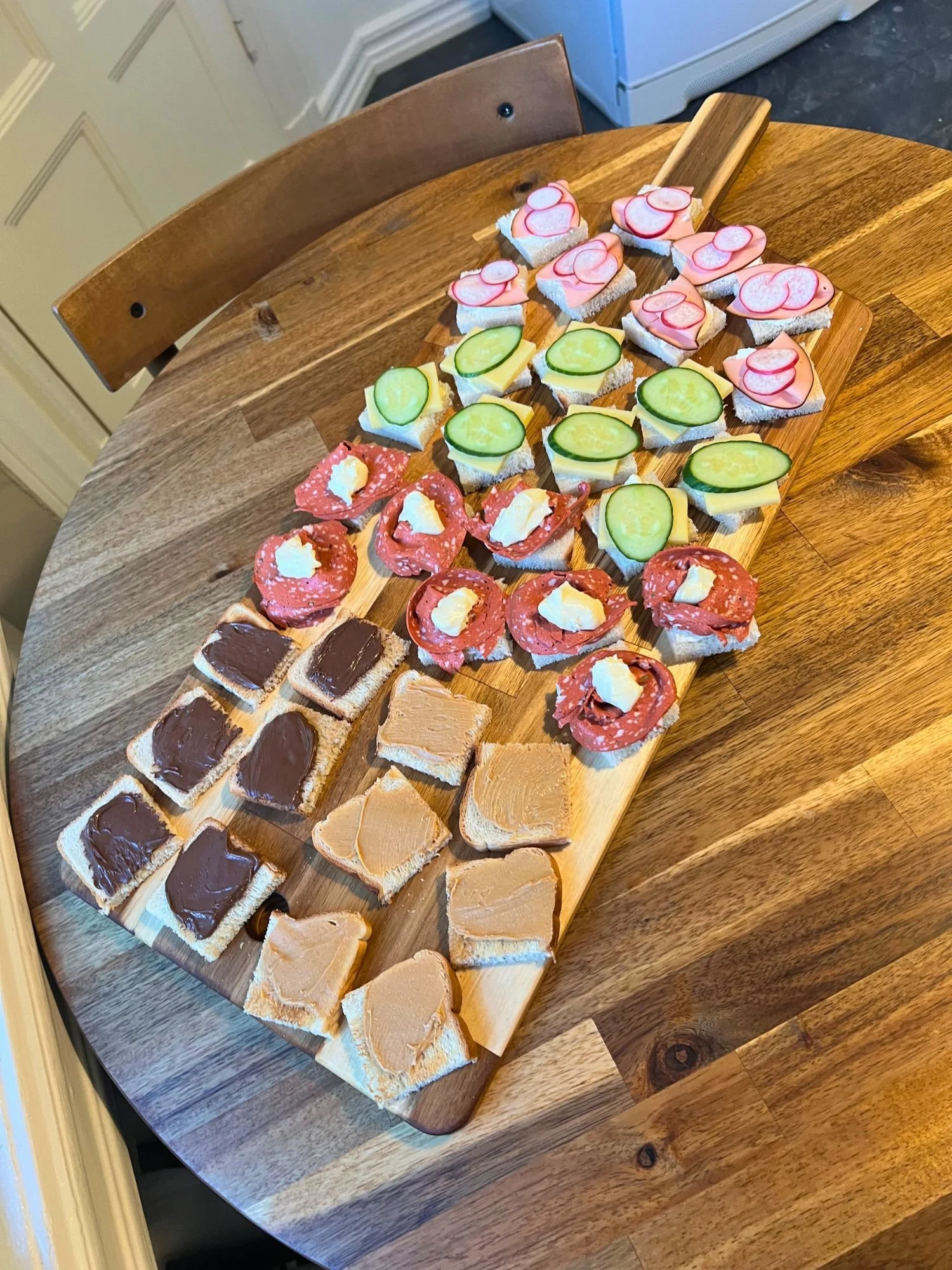 A long wooden serving board rests diagonally across a round wooden table, neatly arranged with rows of bite-sized open-faced mini sandwiches.