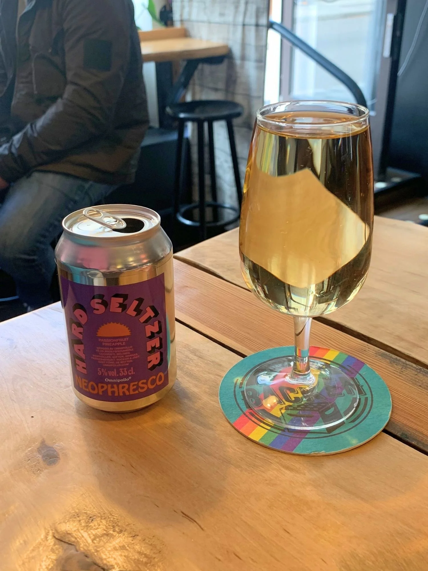 An open beverage can with a purple label that reads "HARD SELTZER NEOPHRESCO" stands on a wooden table next to a stemmed glass filled with a clear, pale yellow drink resting on a colorful, rainbow-bordered coaster.