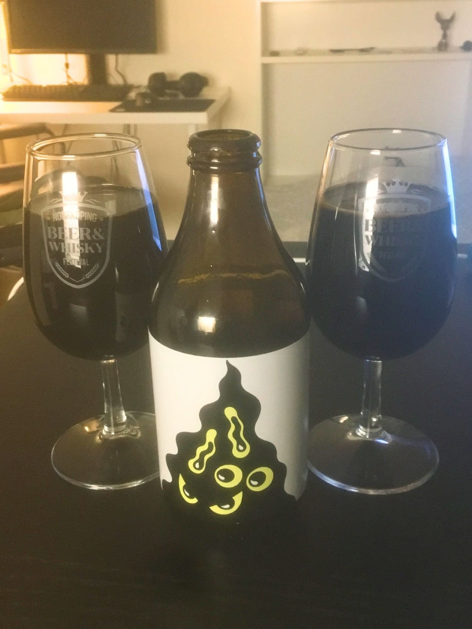 An opened brown beer bottle between two glasses filled with dark imperial stout on a dark brown table. The bottle features a white label with a kokostopp graphic illustrated with yellow eyes and abstract squiggles.