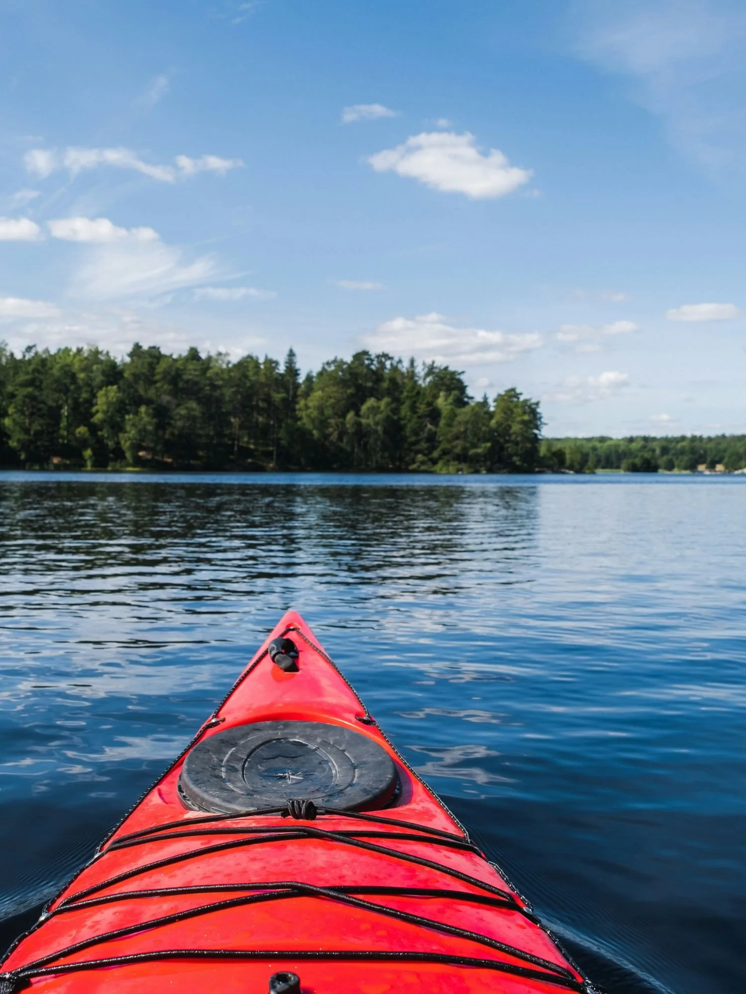 The bow of a bright red kayak as it glides across a calm, blue lake.  The front of the kayak features a black circular hatch and bungee cords stretched across the deck. Small ripples fan out from the boat into the deep blue water.