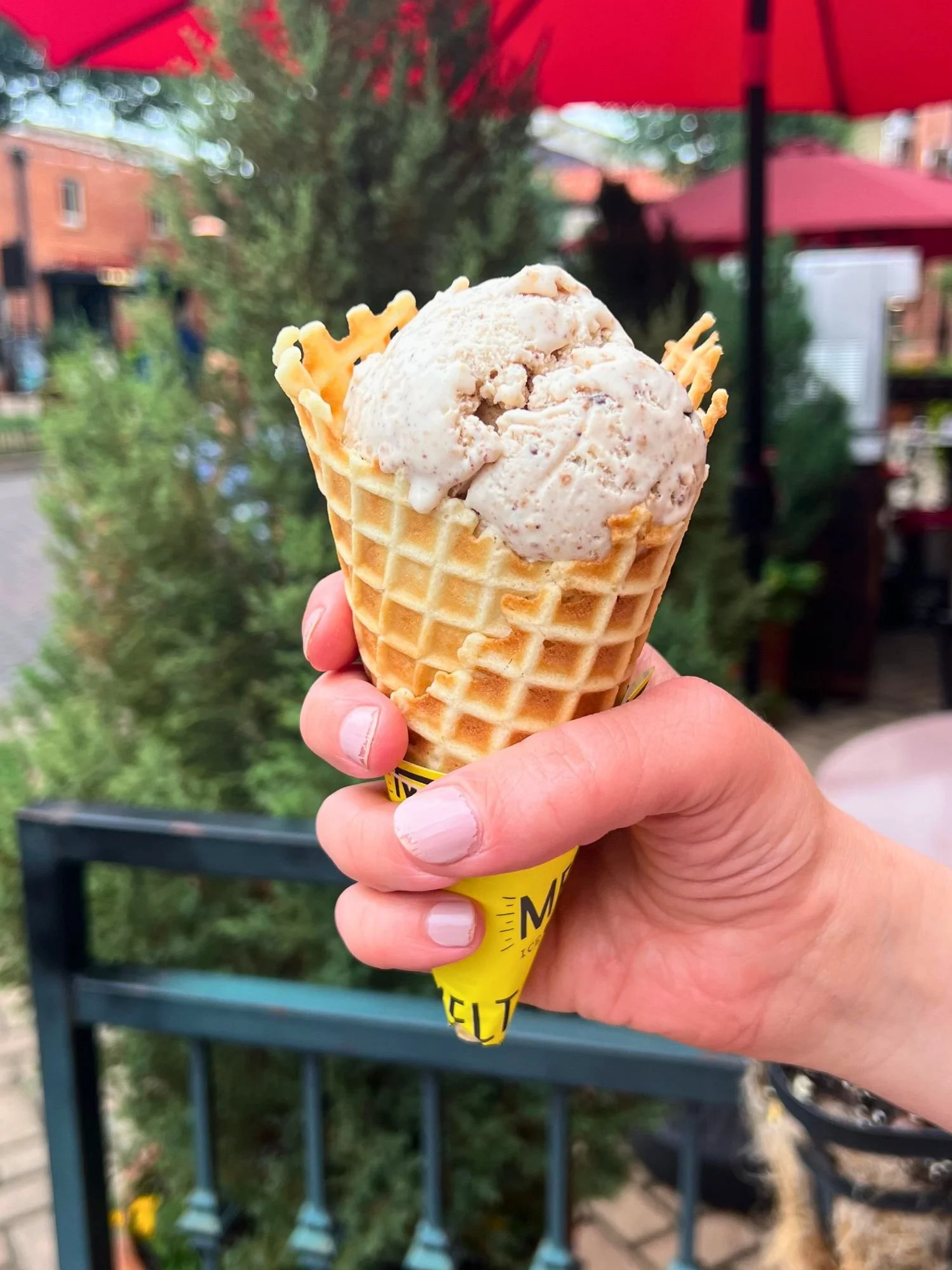 A hand holding a large scoop of light brown ice cream with dark flecks in a waffle cone wrapped in a yellow paper sleeve. The background is a blurry outdoor patio with green trees and red umbrellas.