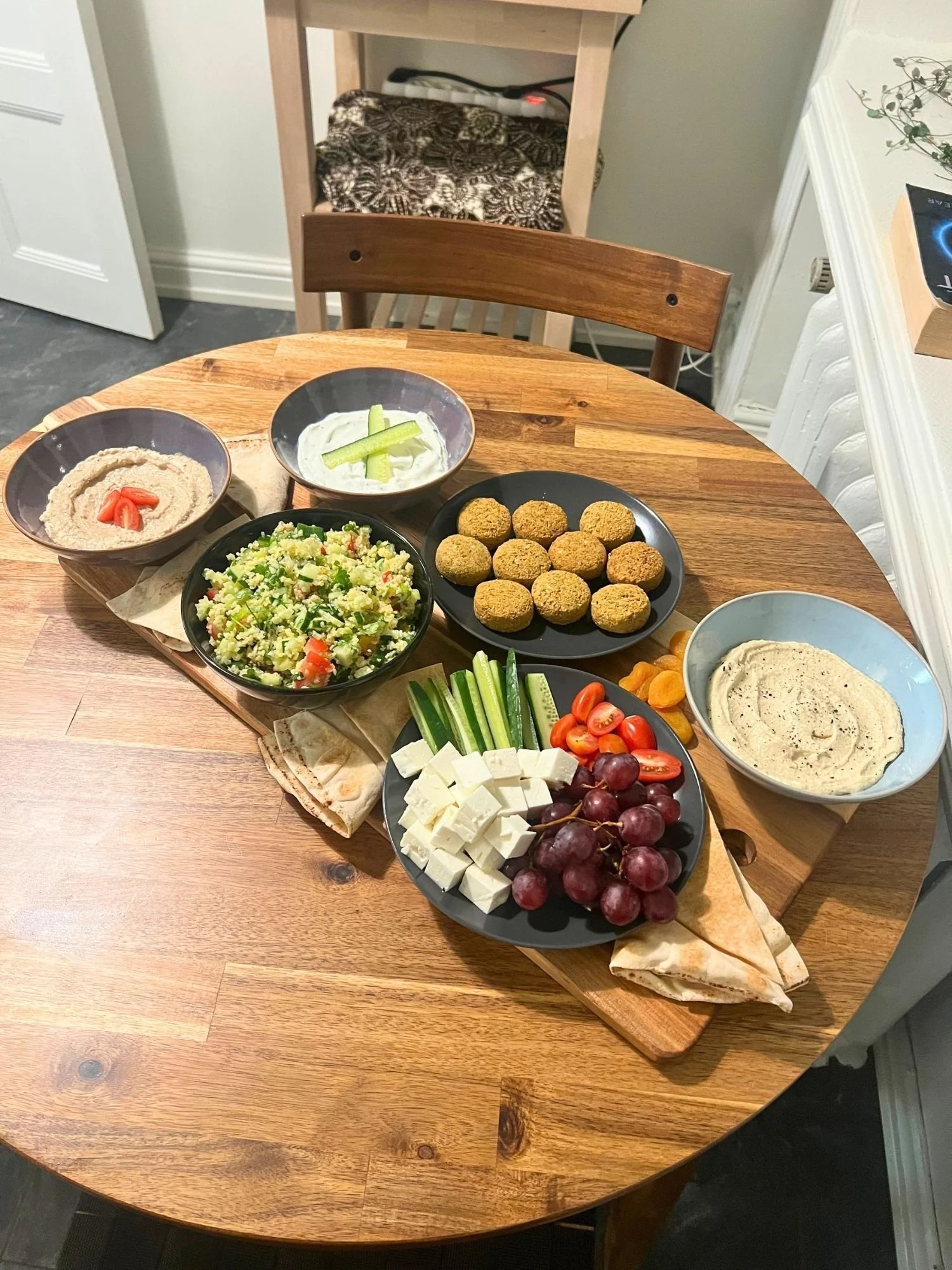 A wooden serving board on a round wooden table filled with a Mediterranean mezze spread, including falafel, tabbouleh salad, dips, feta cheese, fresh vegetables, red grapes, and pita bread.