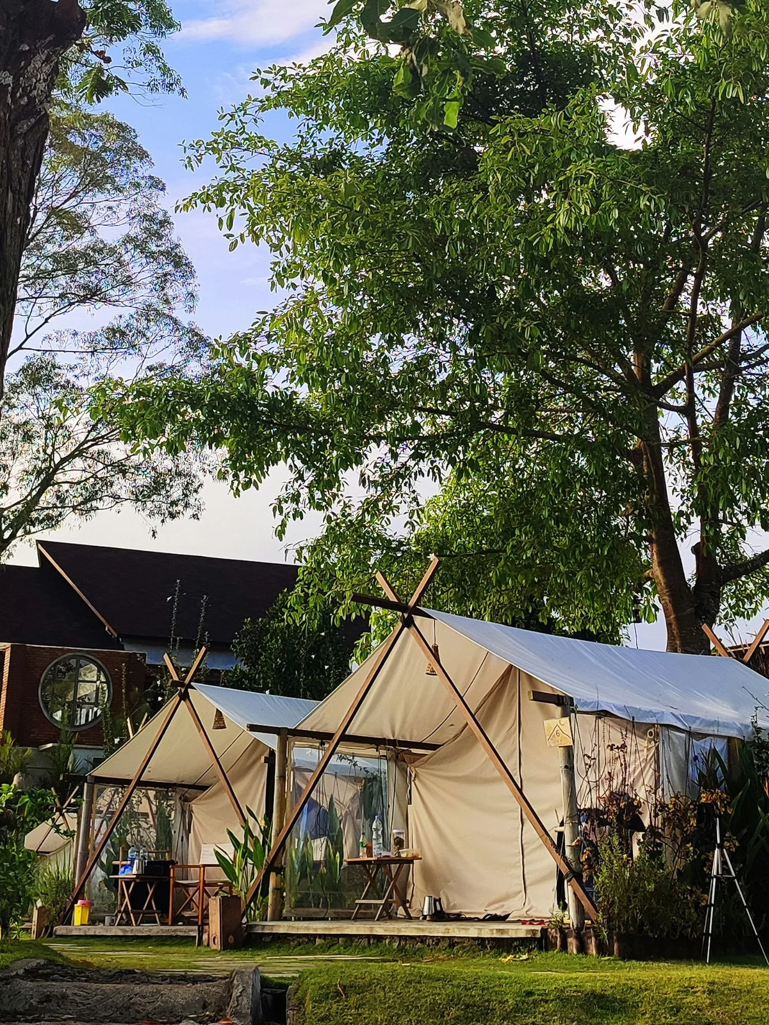 Two large, white safari-style glamping tents are pitched side-by-side on raised wooden platforms on a green lawn. Each tent features a distinctive external wooden A-frame structure supporting a white canopy over the entrance.