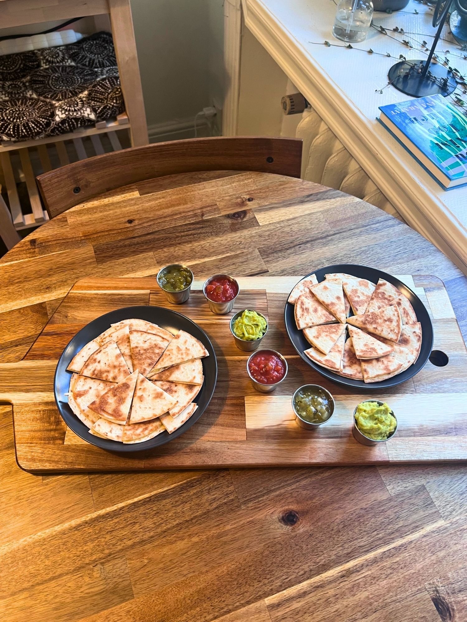 A wooden serving board on a round wooden table, arranged with two dark plates of sliced quesadilla triangles and small ramekins of salsa and guacamole.
