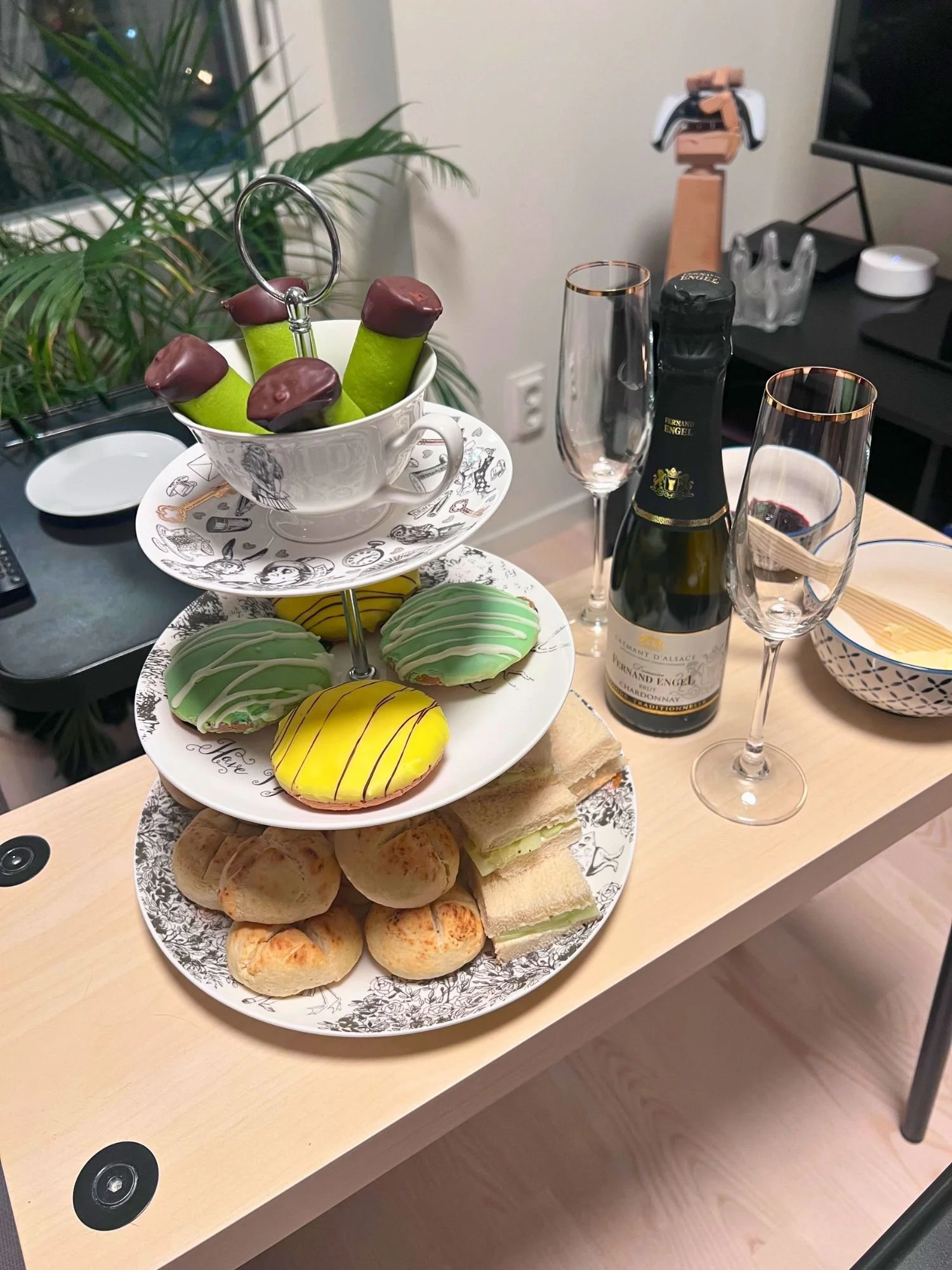 An afternoon tea spread on a light wood table. The top tier has a white teacup with black illustrations with four green dammsugare pastries with chocolate-dipped ends.
