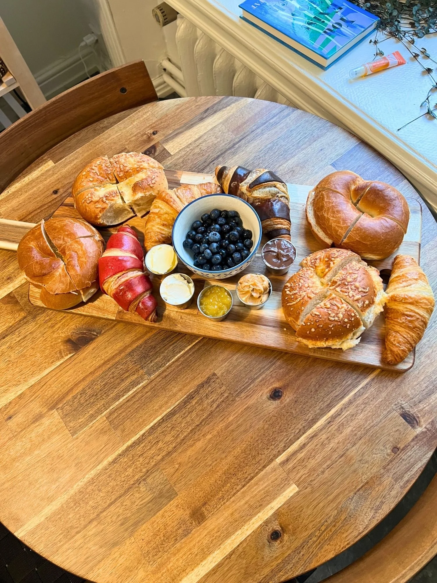 A long wooden serving board rests on a round wooden table, filled with a breakfast spread of bagels, croissants, and toppings.